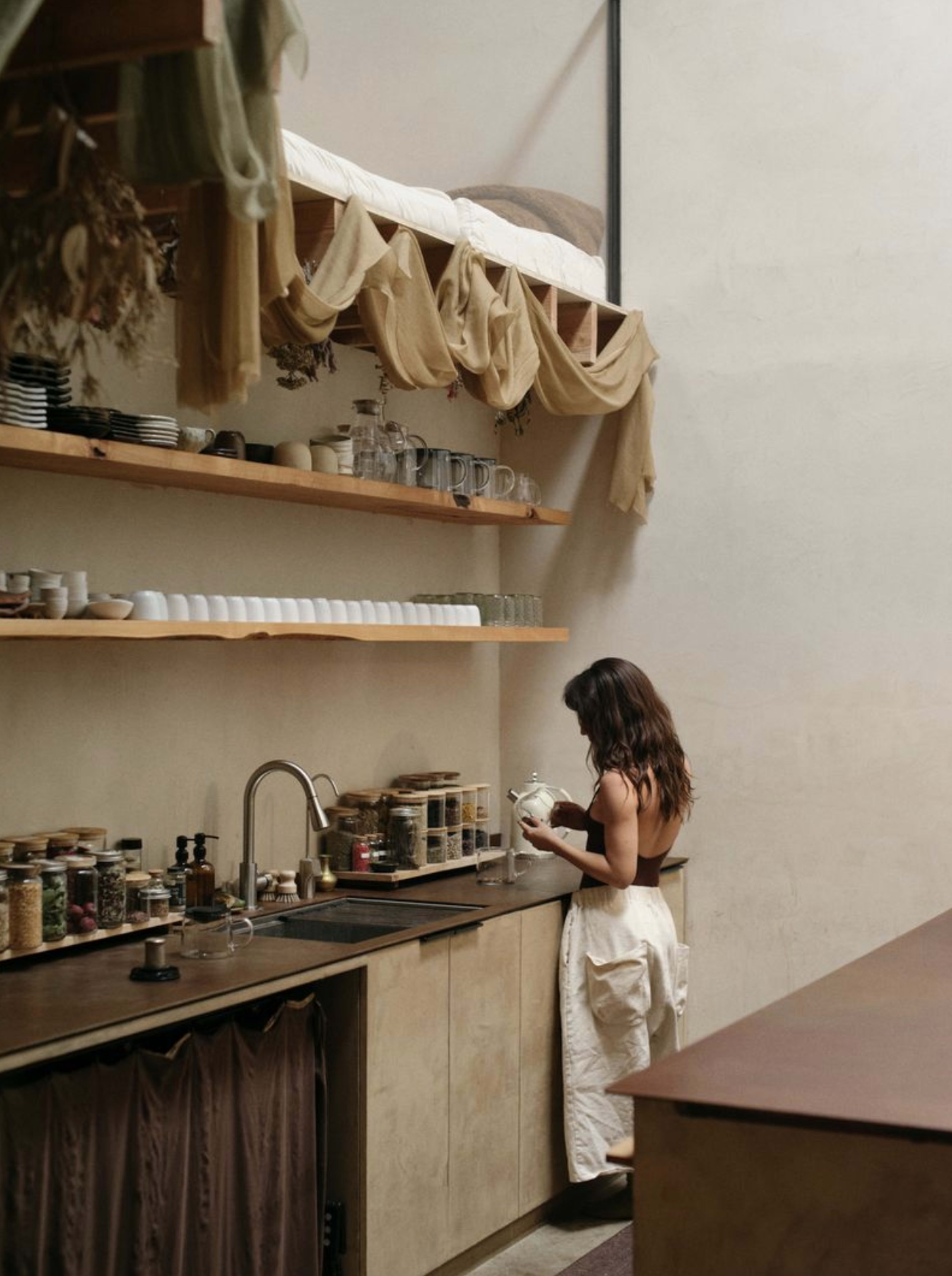 A woman stands at a kitchen sink in a minimalist space, surrounded by shelves filled with jars and dishes, while a bed is positioned above her.