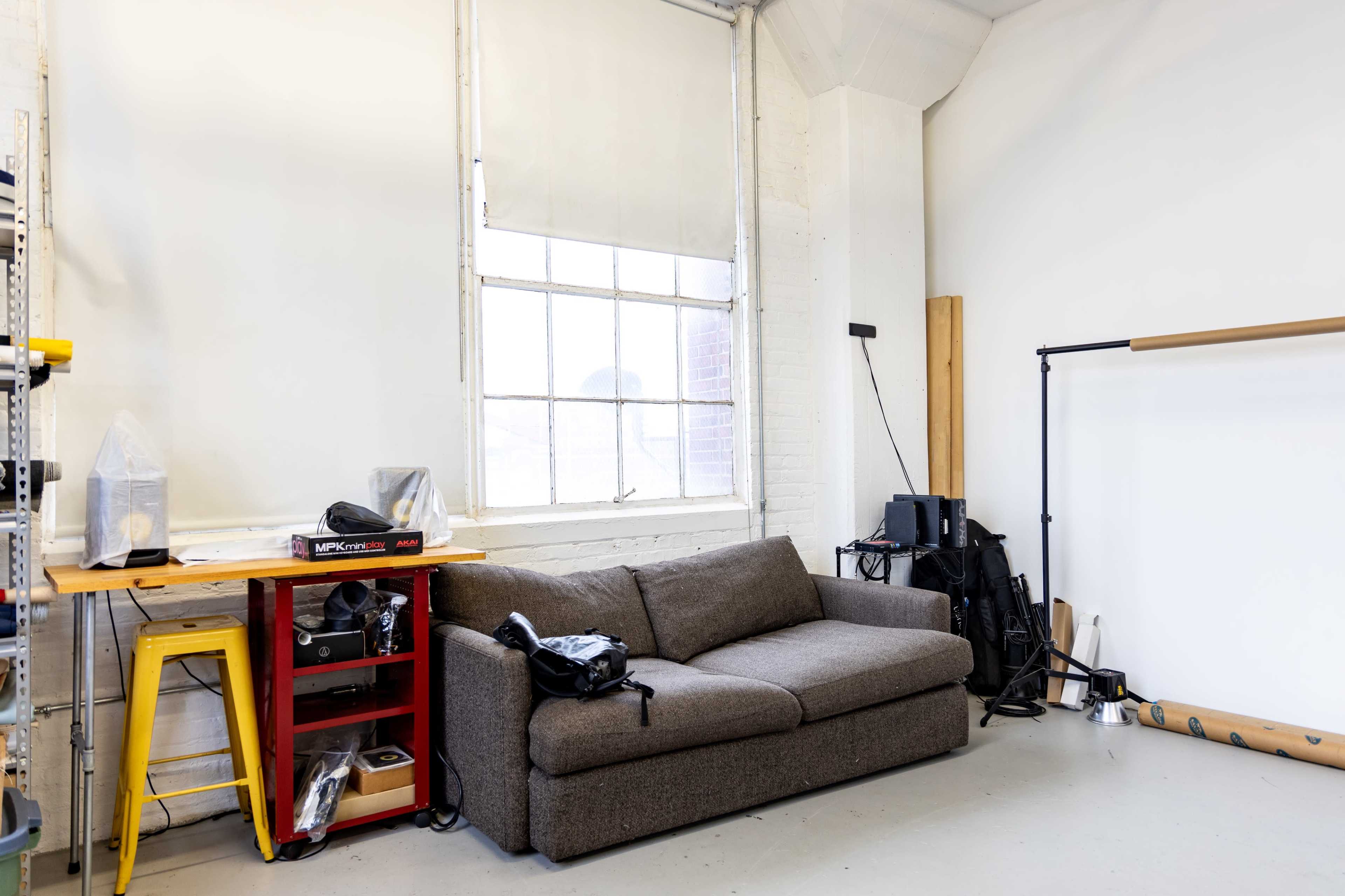 A gray couch is placed against a bare wall in a bright room with a large window and a worktable beside it.