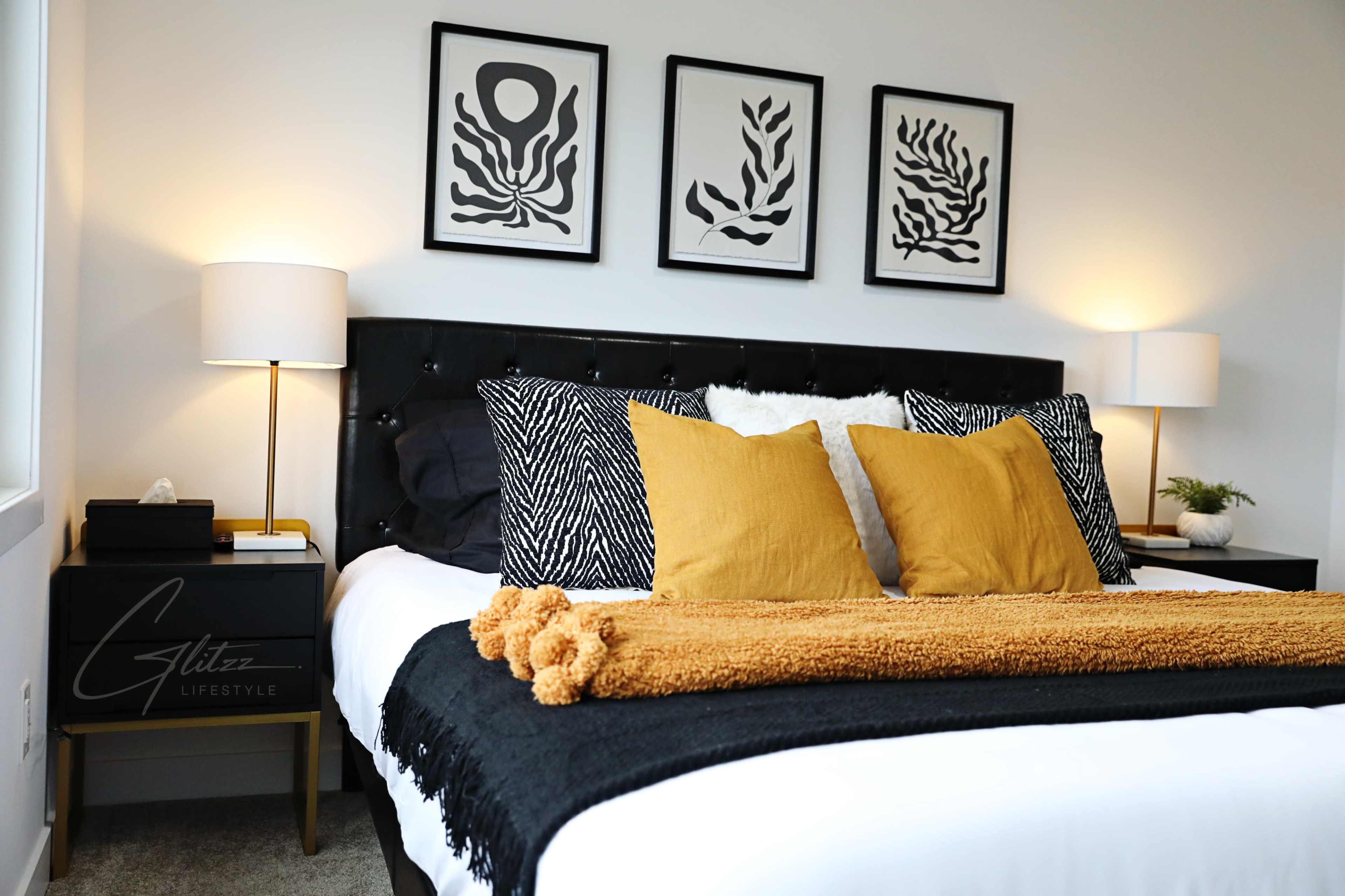 A modern bedroom featuring a black upholstered bed with decorative pillows and a mustard-colored throw, flanked by bedside tables with lamps and framed wall art.
