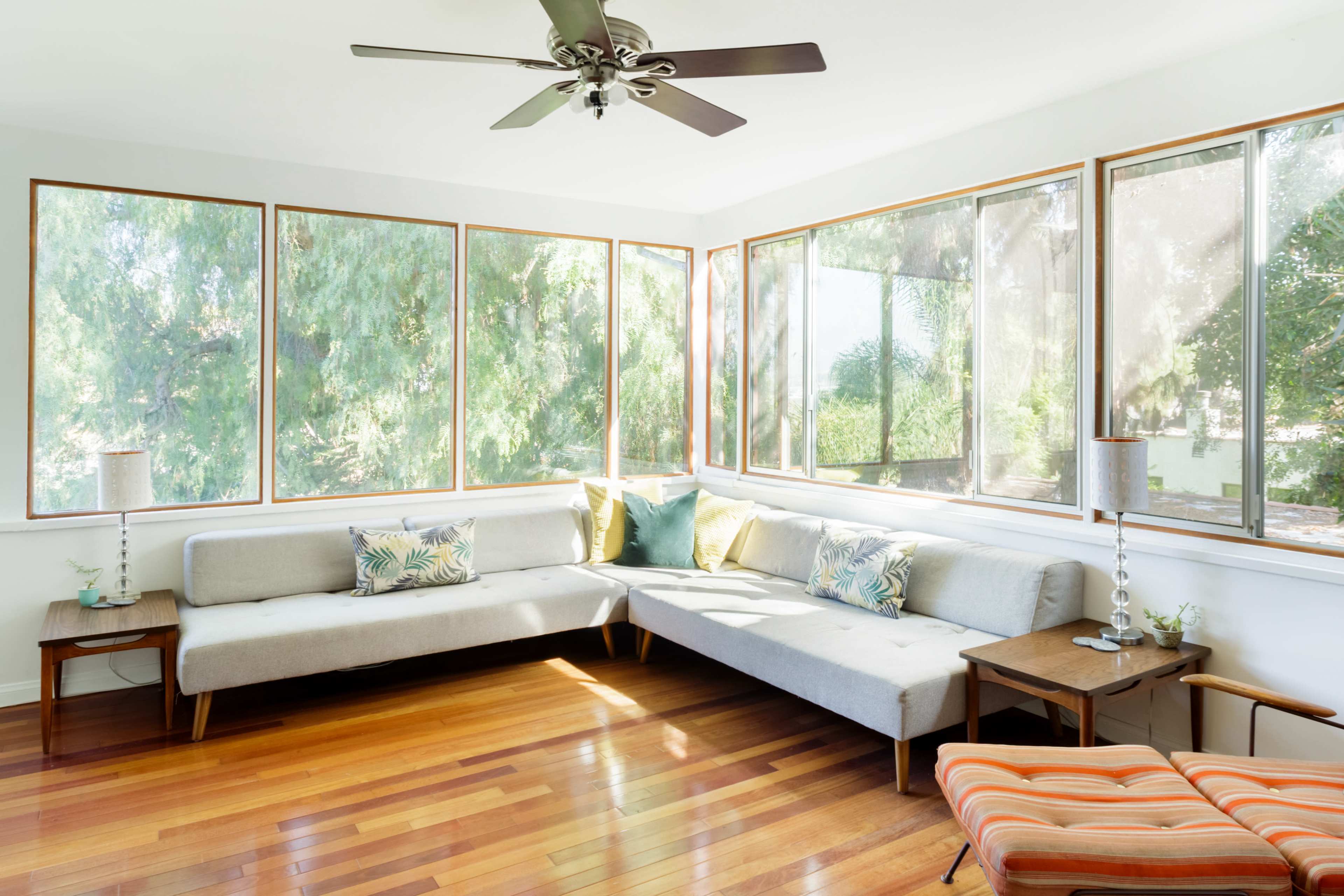 A sunlit living room features a beige sectional sofa, wooden floors, and large windows with views of greenery.