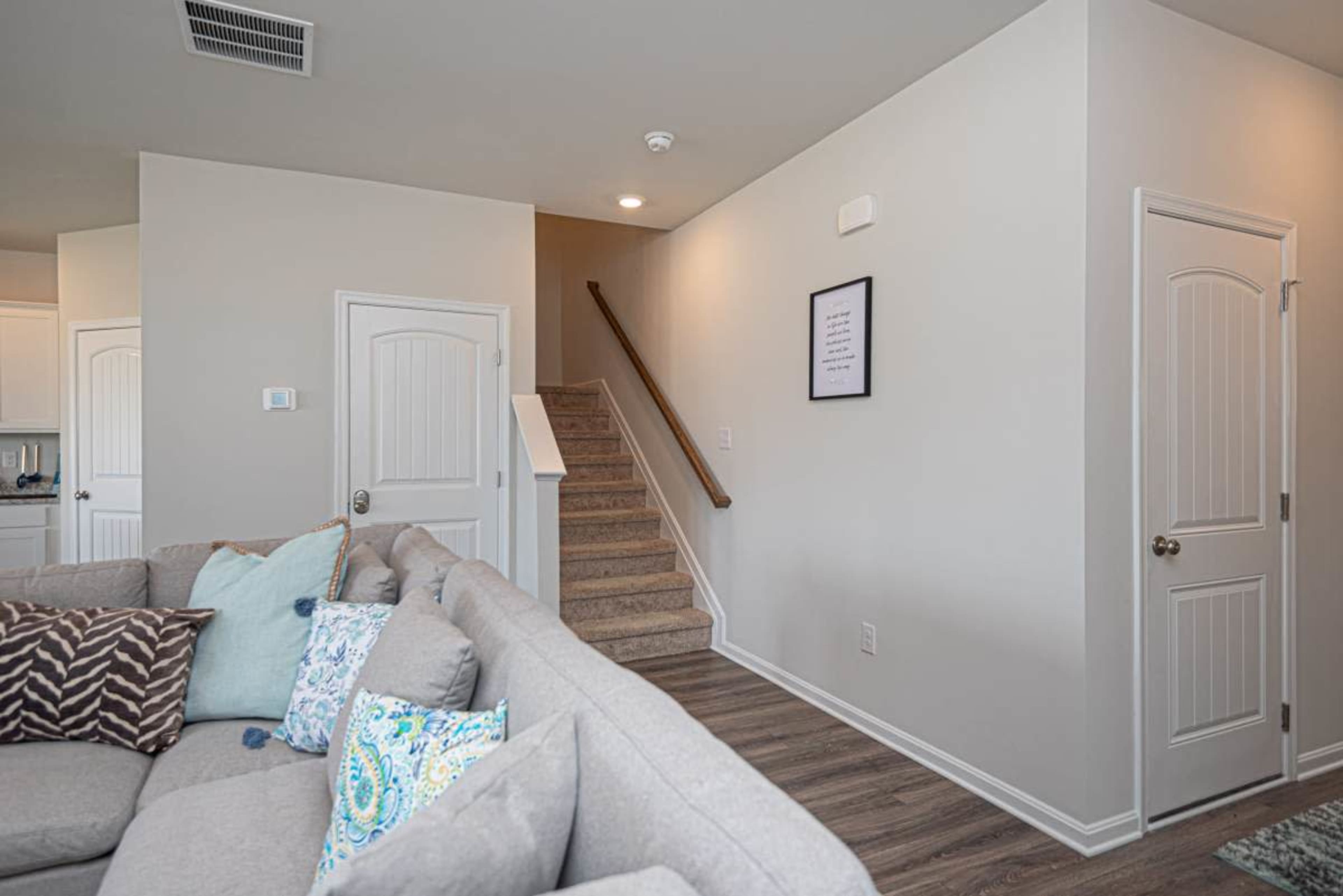 A living room with a grey sectional sofa, a staircase in the background, and a door leading to another room.