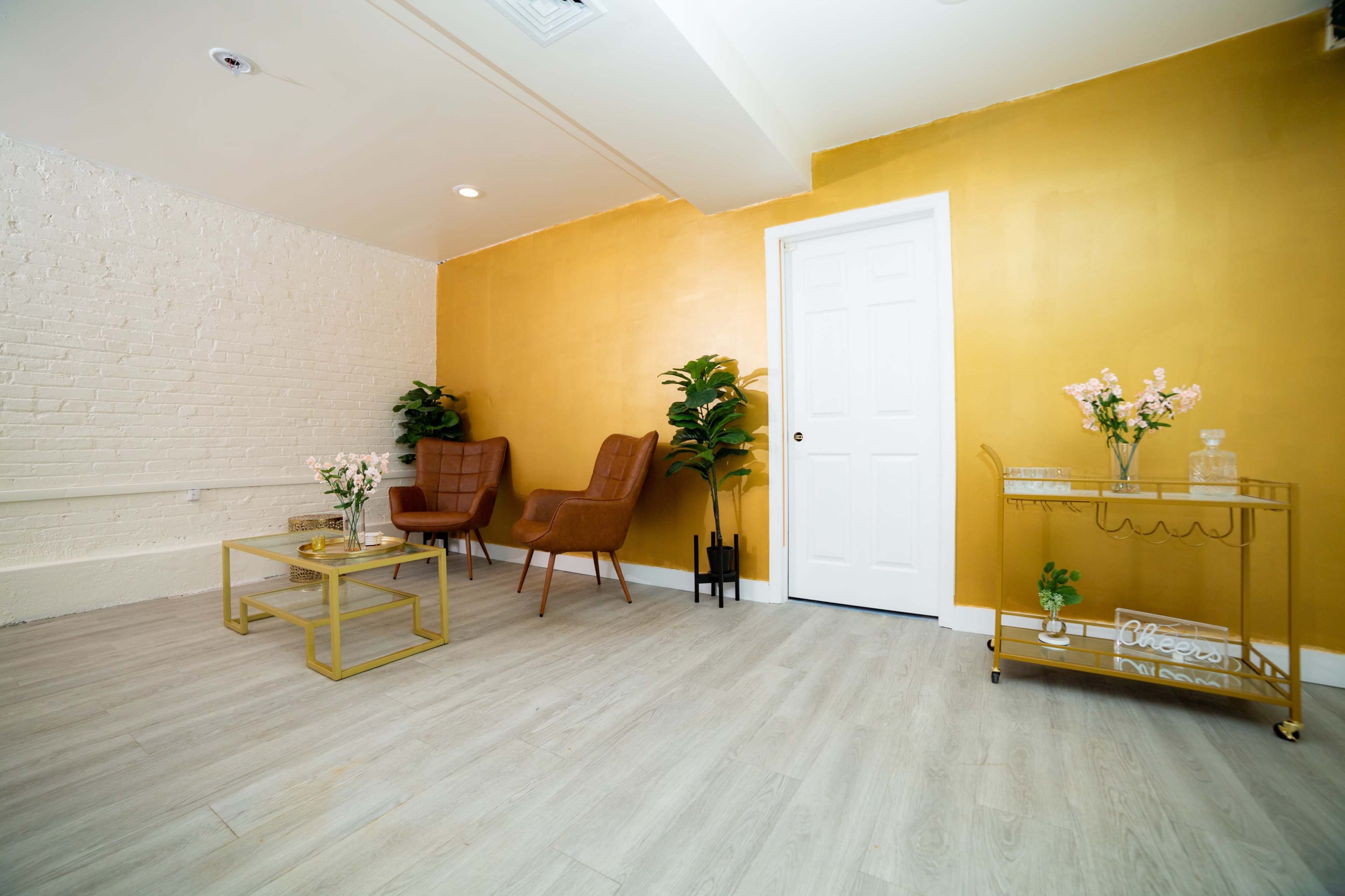 A room with a golden yellow accent wall, two brown armchairs, a small coffee table, and a decorative cart with plants and flowers.