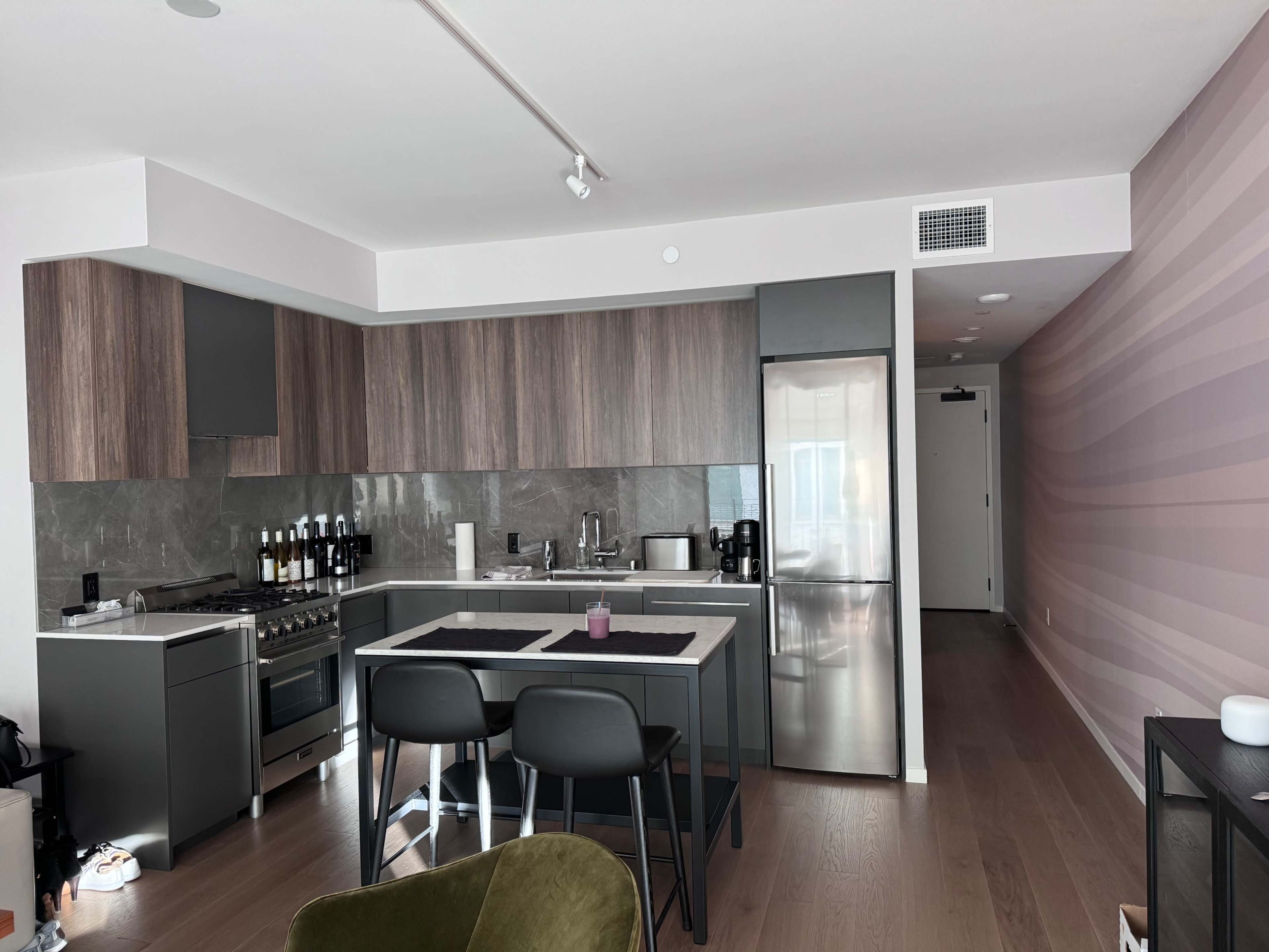 A modern kitchen with wooden cabinetry, stainless steel appliances, and a small dining area with two black stools, adjacent to a hallway.