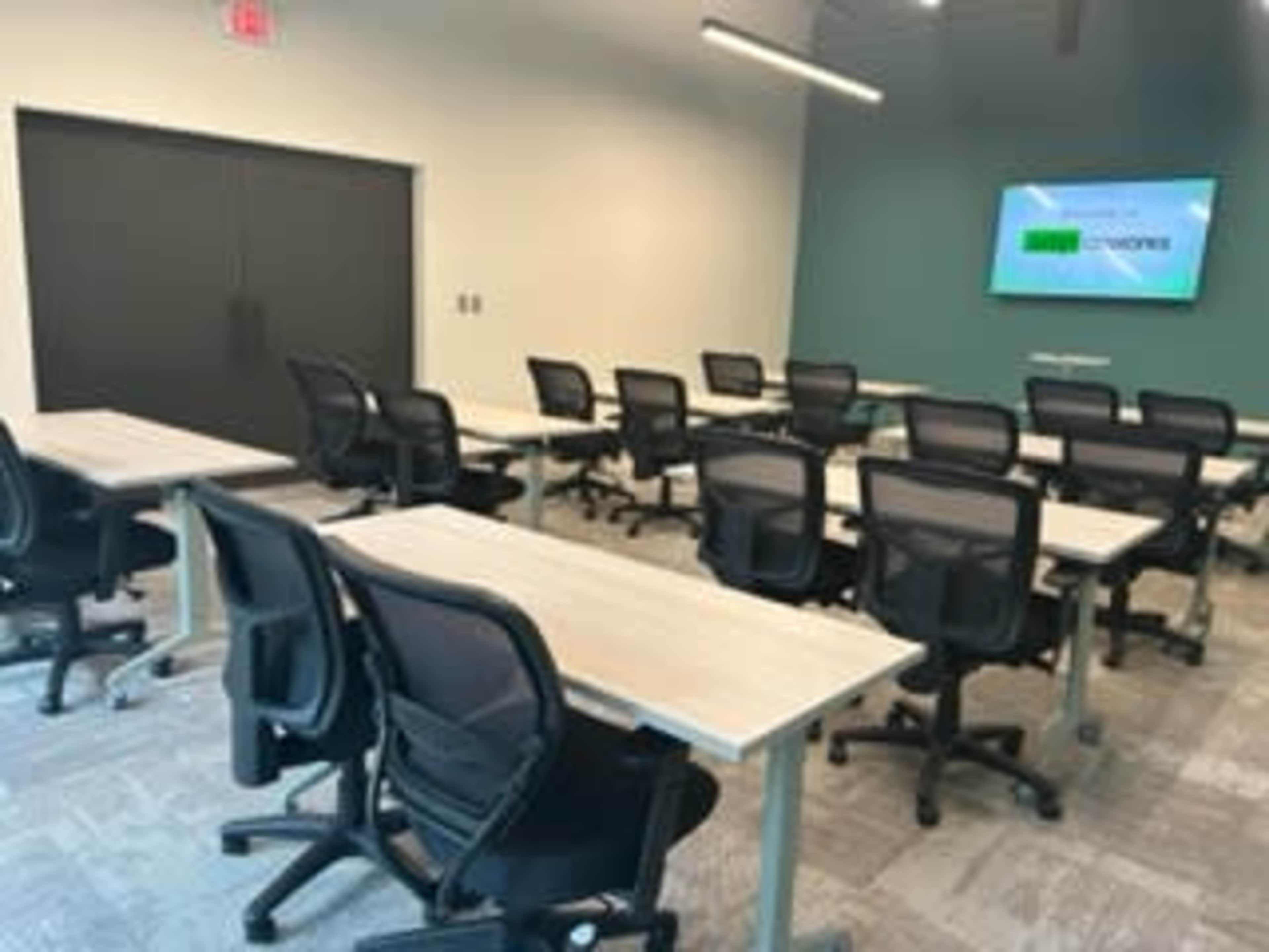 A modern classroom or meeting room with multiple tables and ergonomic chairs arranged in rows, featuring a large screen on one wall.