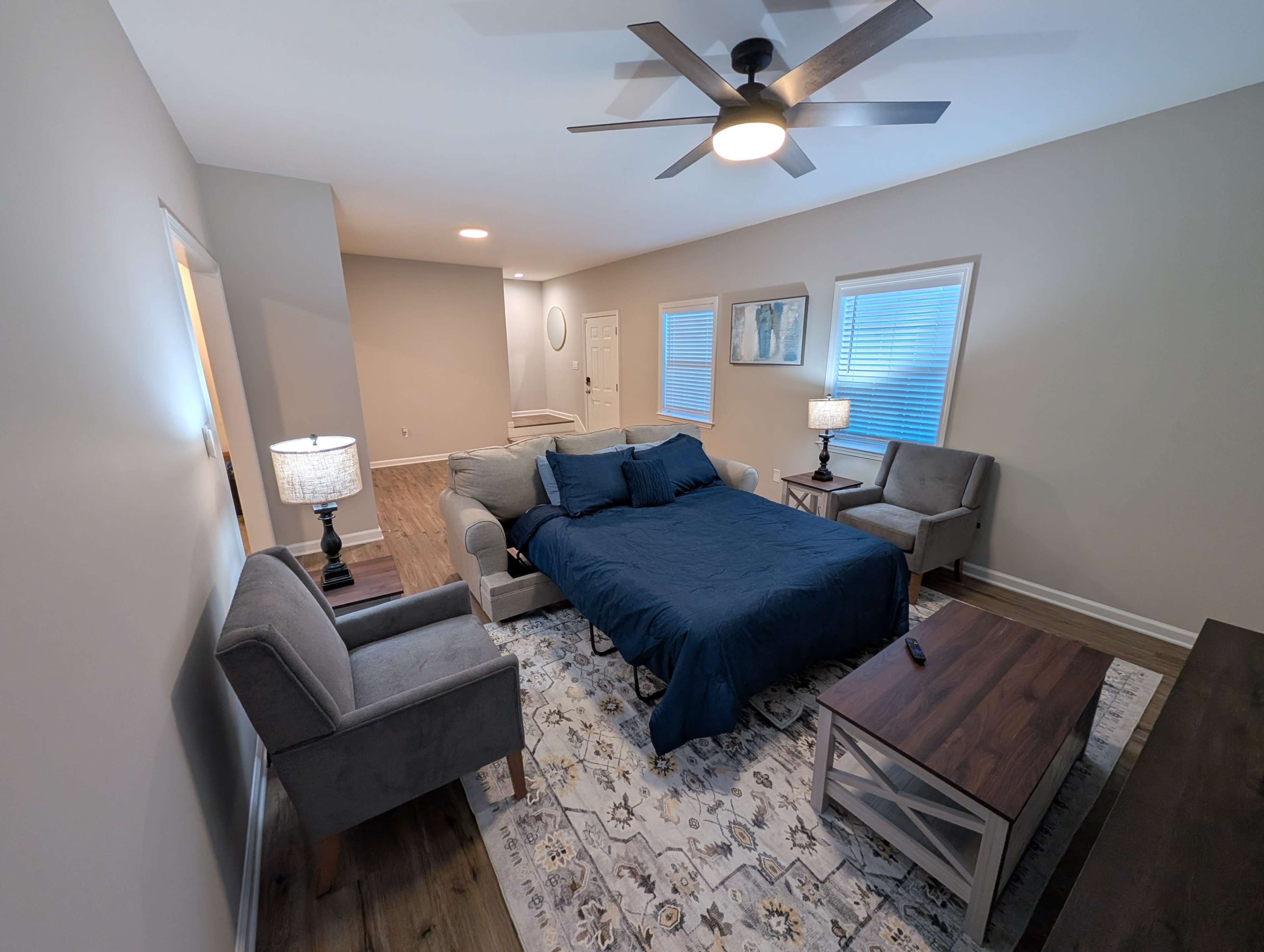 The image shows a spacious bedroom featuring a large bed with a blue bedspread, two armchairs, a coffee table, and a lamp on each side of the bed, all arranged against neutral-colored walls.