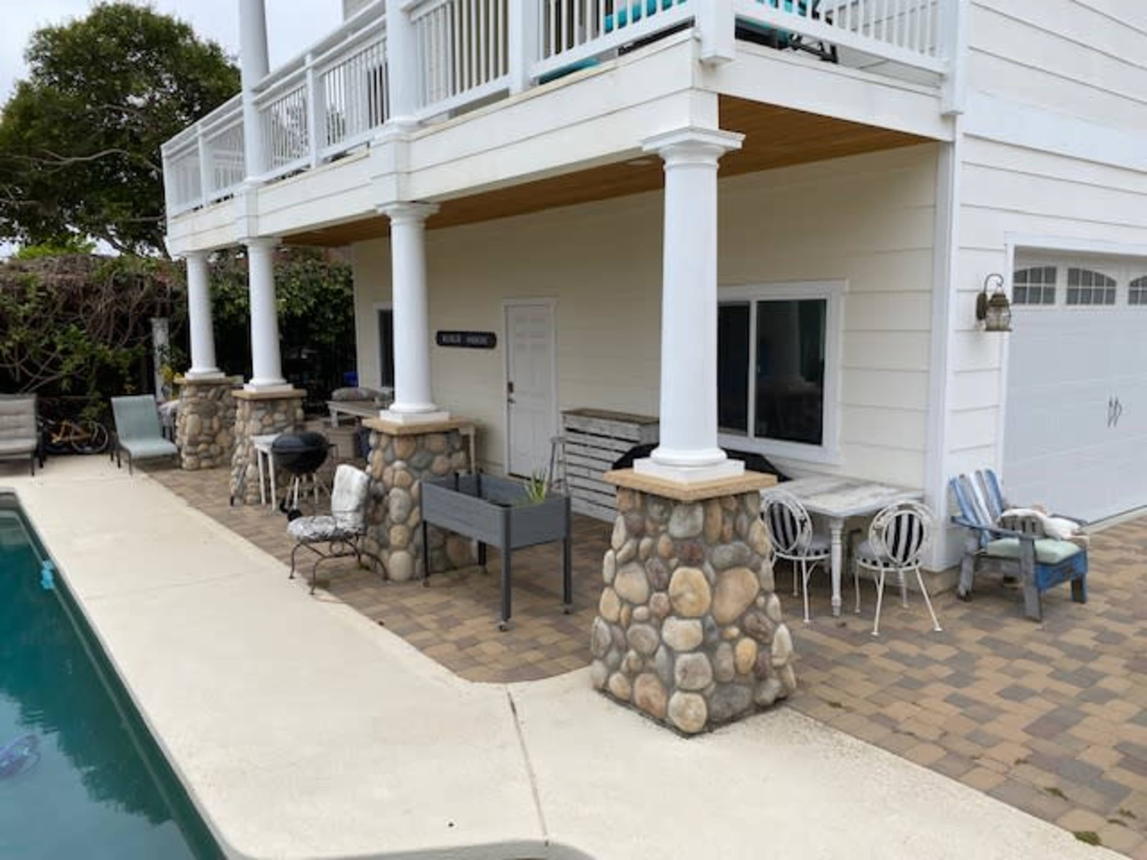 The image shows a patio area beside a swimming pool, featuring a house with a light-colored exterior, stone column bases, and some outdoor furniture.