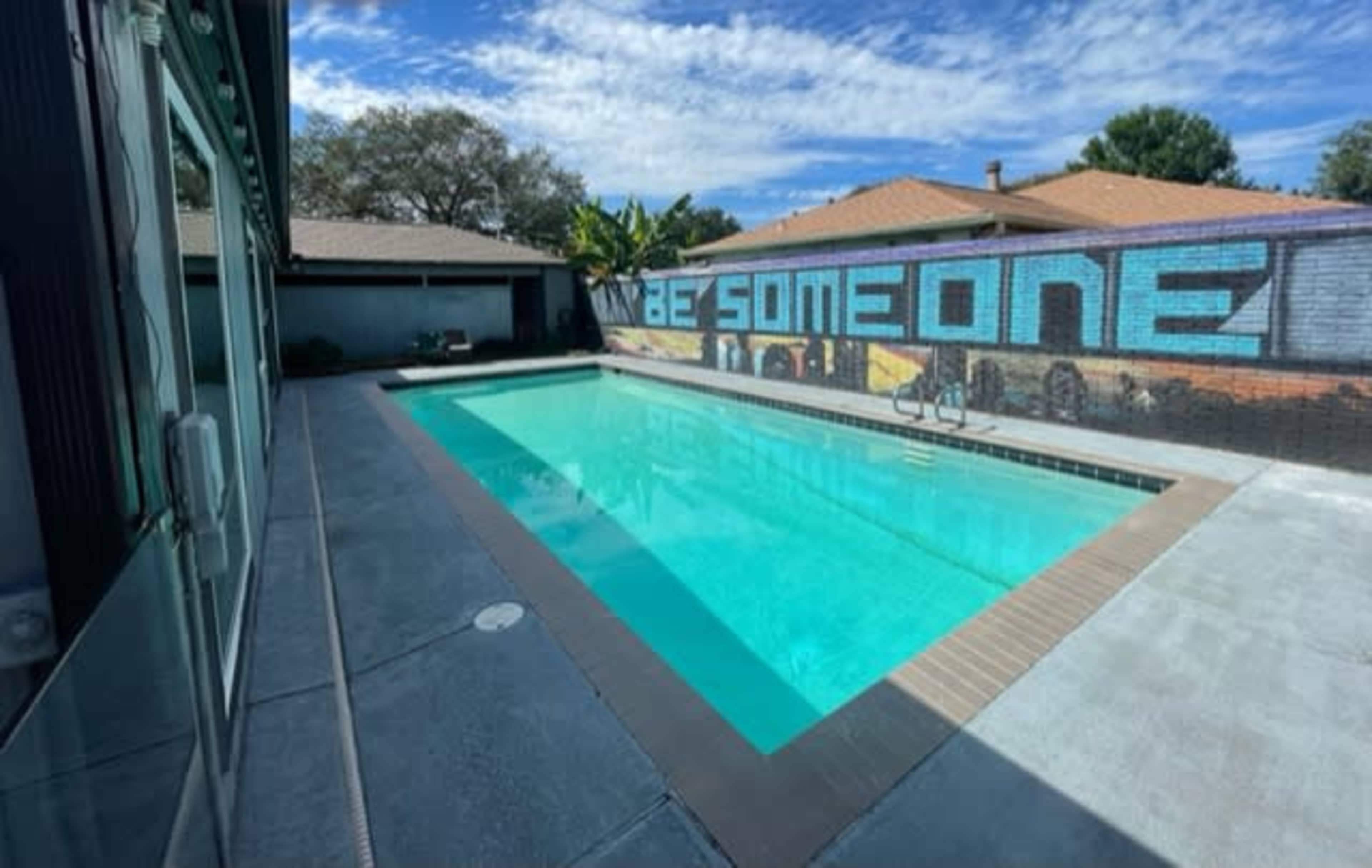 A clear swimming pool is situated alongside a mural that reads "BE SOMEONE," with houses and trees visible in the background.