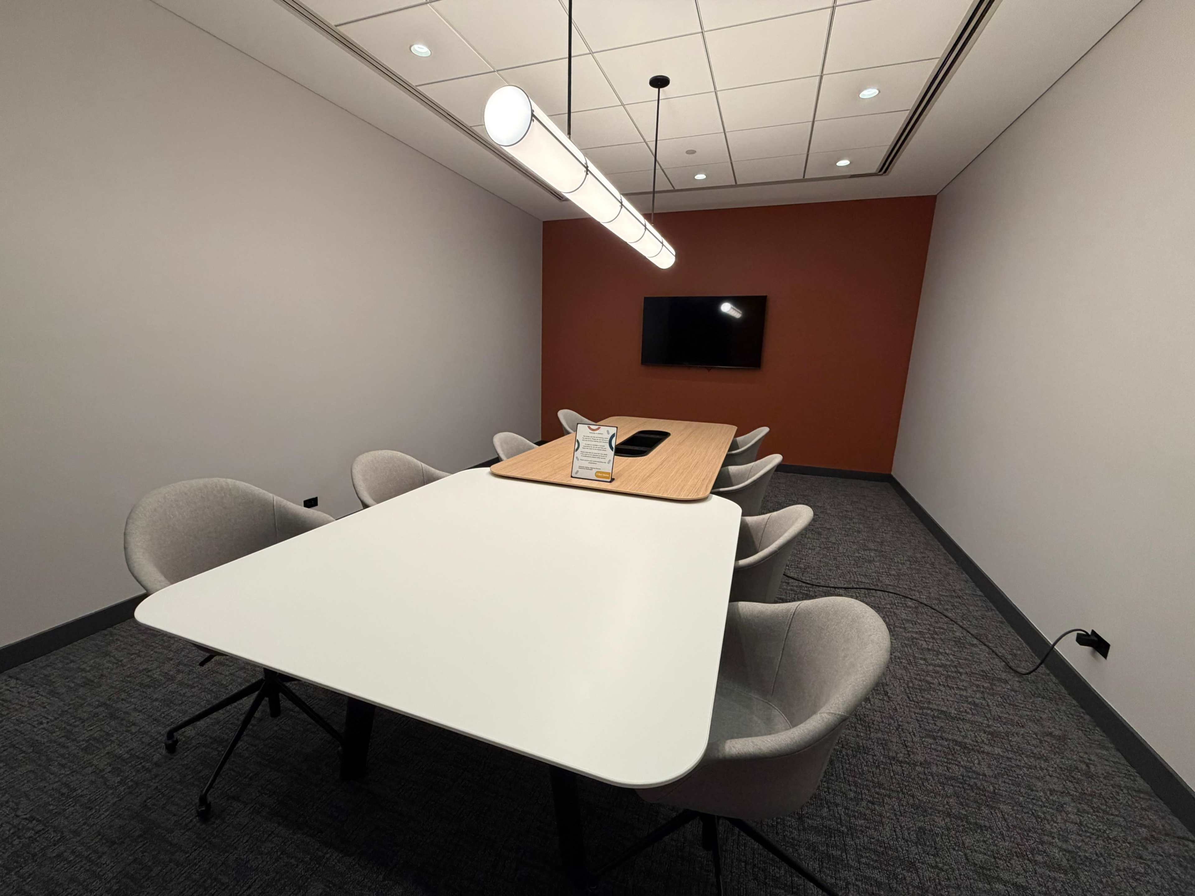 A modern conference room features a long, light wood table surrounded by gray chairs, with a wall-mounted television and pendant lighting above.