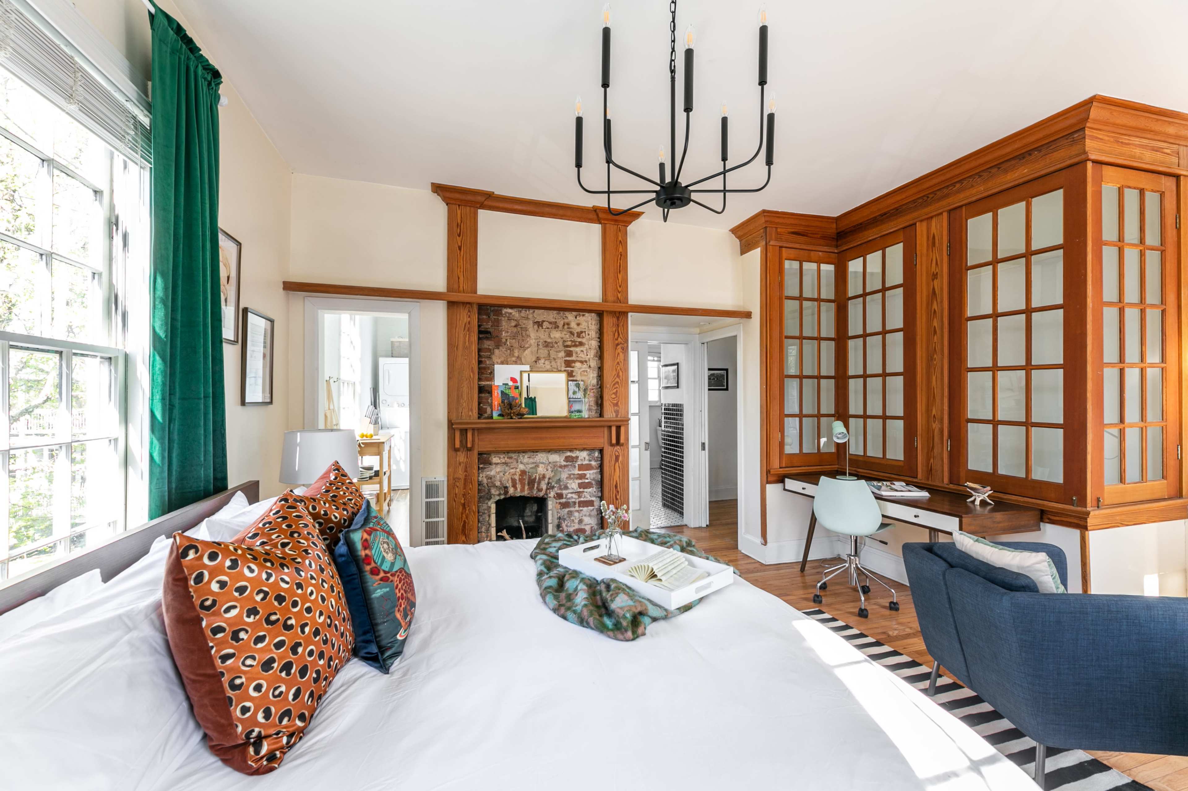 A well-lit bedroom featuring a large bed with decorative pillows, a wooden desk and chair against a wall with glass panels, and a fireplace surrounded by exposed brick.