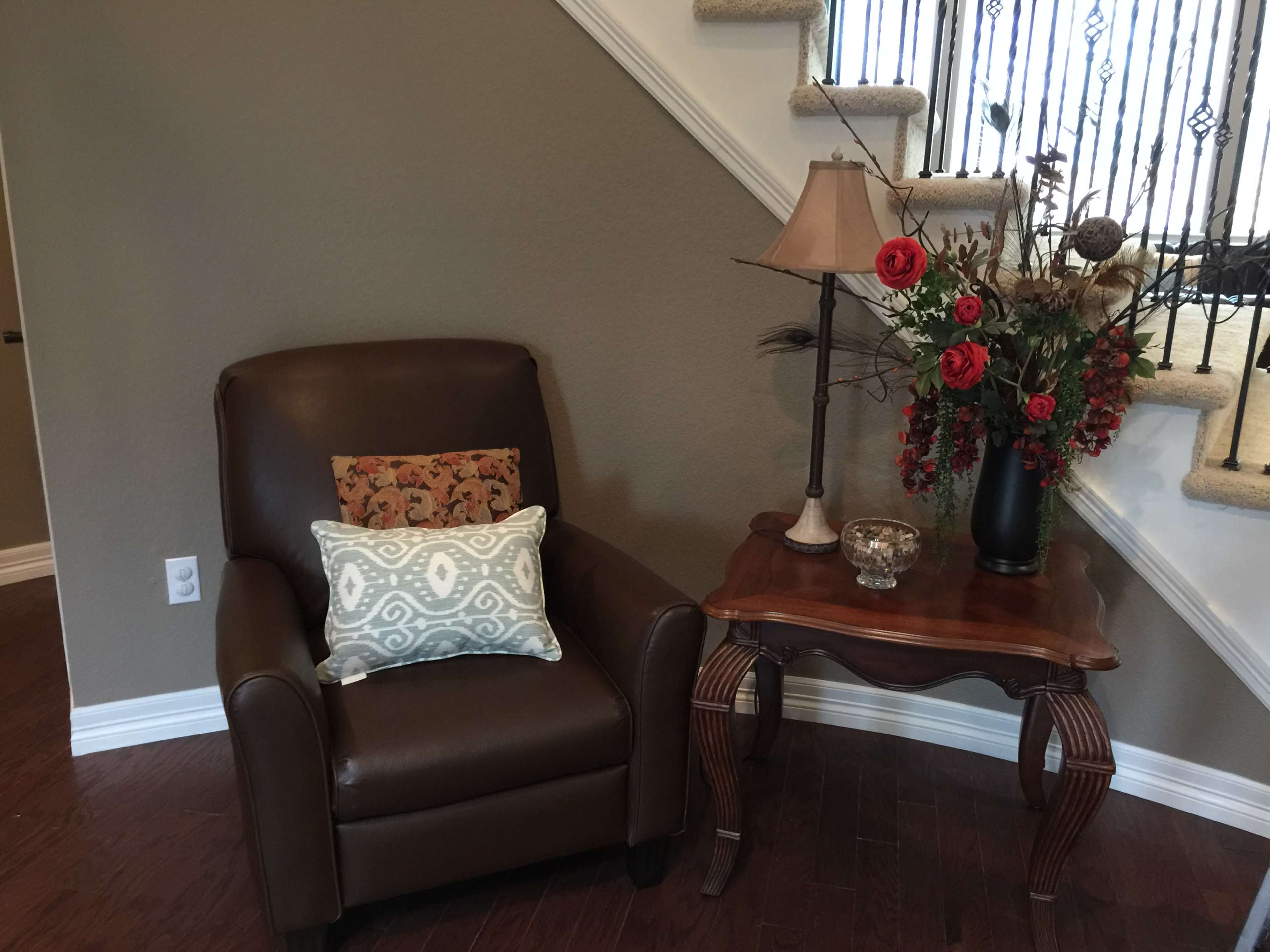 A brown armchair with decorative pillows sits beside a wooden side table topped with a lamp and a vase of flowers in a well-lit corner near a staircase.