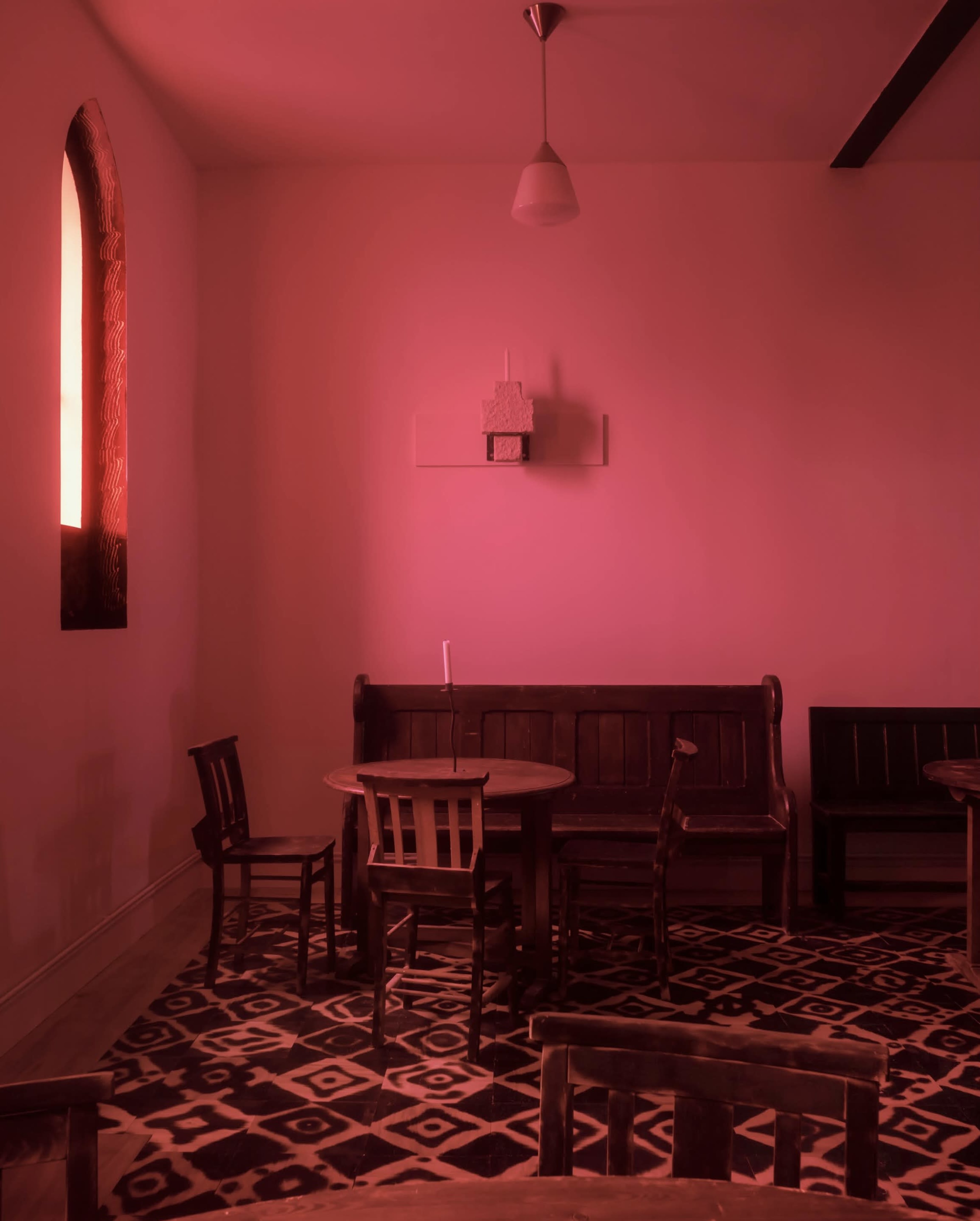 A small room features a round table surrounded by wooden chairs, illuminated by red light from a window.