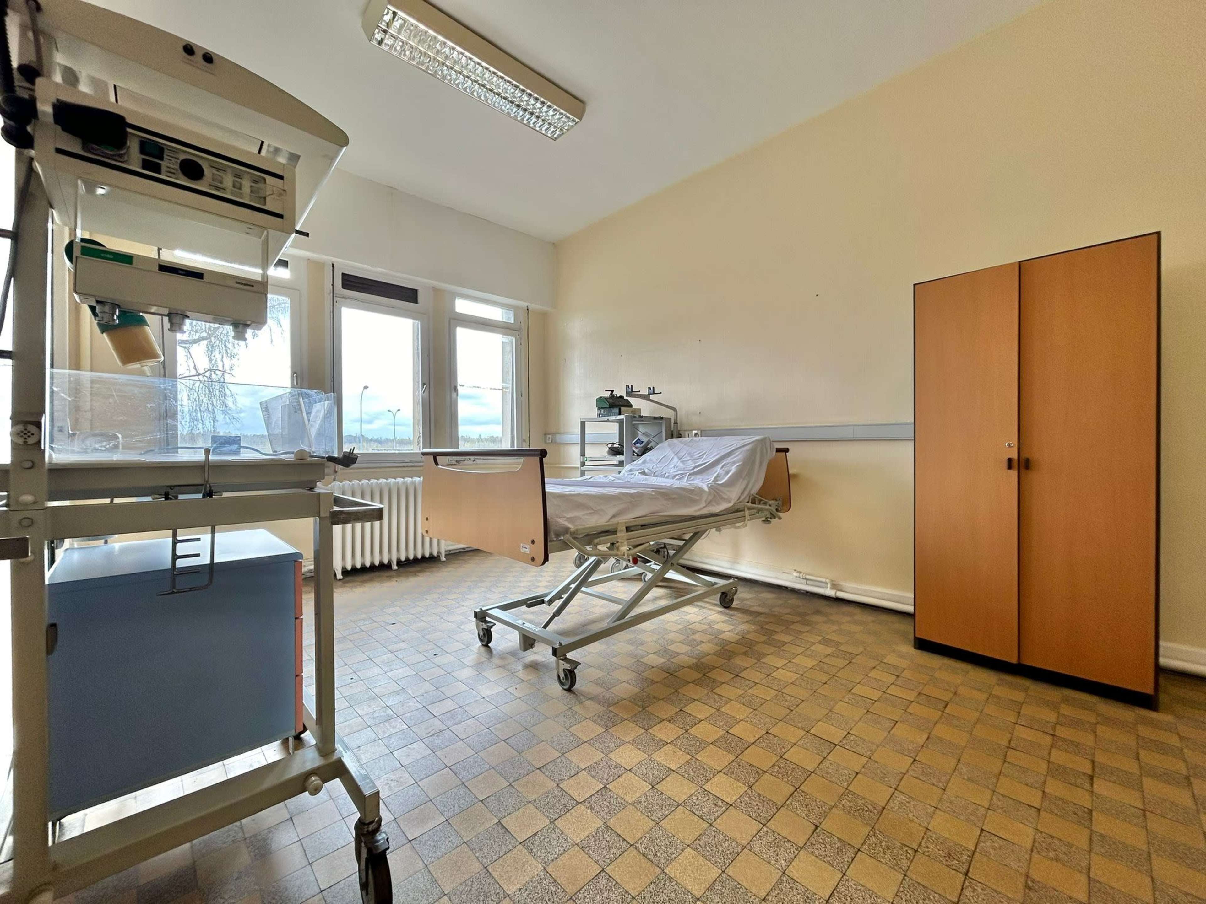 A sparsely furnished hospital room with a hospital bed, a bedside cart, and a wardrobe.