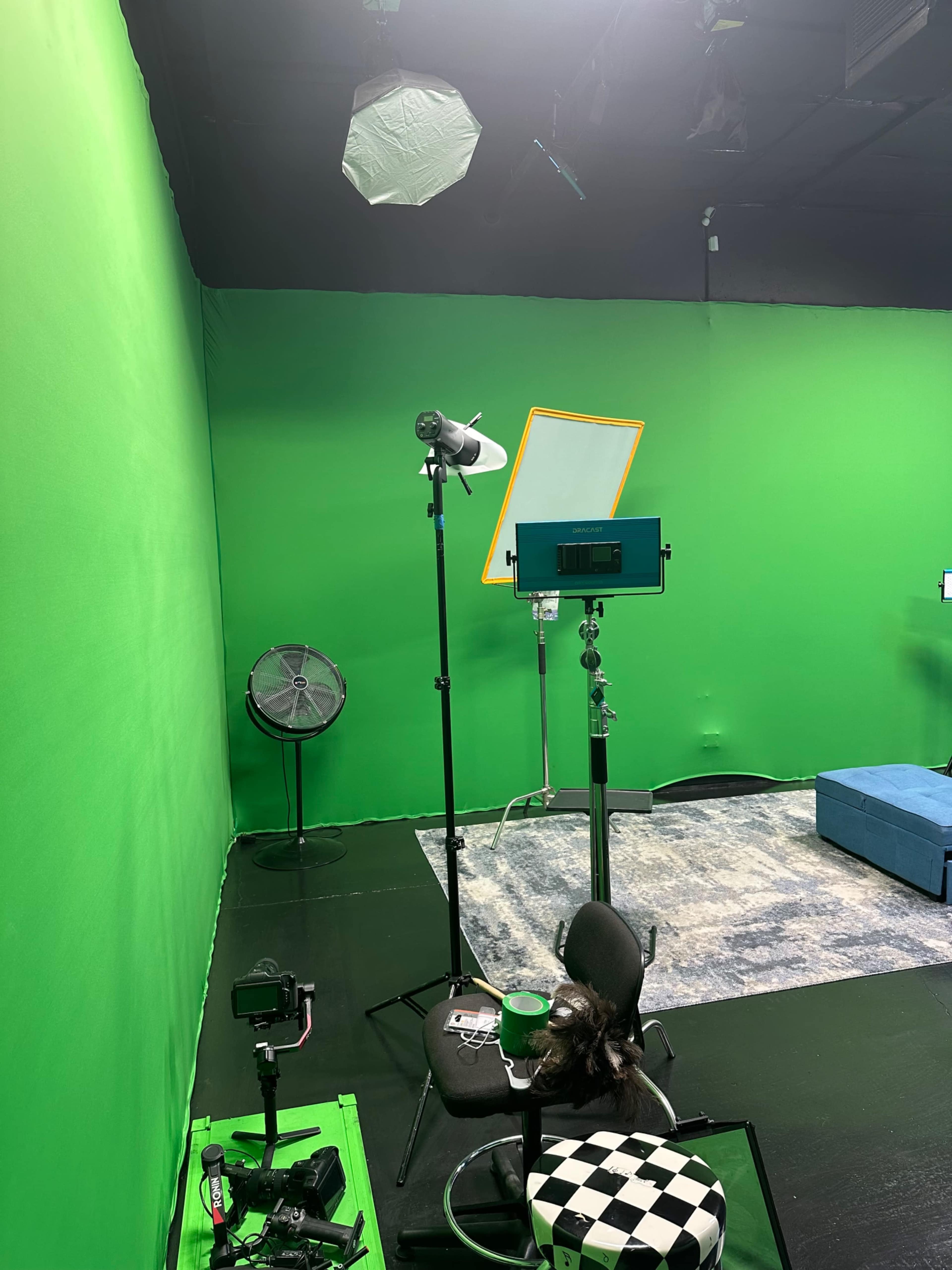 The image shows a green screen studio with lighting equipment, a fan, and a chair on a patterned rug.