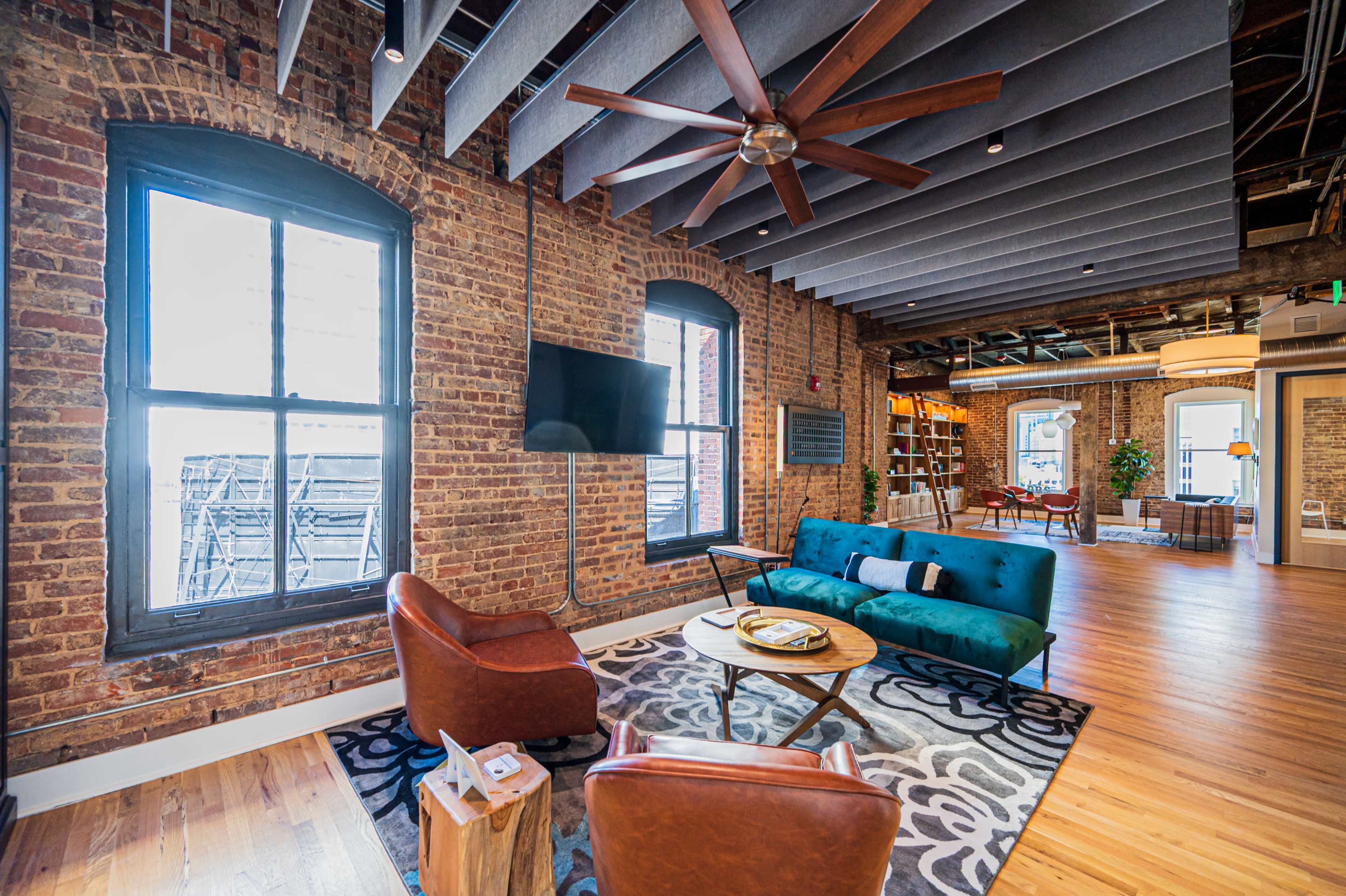 A room featuring exposed brick walls, a blue sofa, two brown leather chairs, a round coffee table, and a ceiling fan, with large windows allowing natural light to enter.