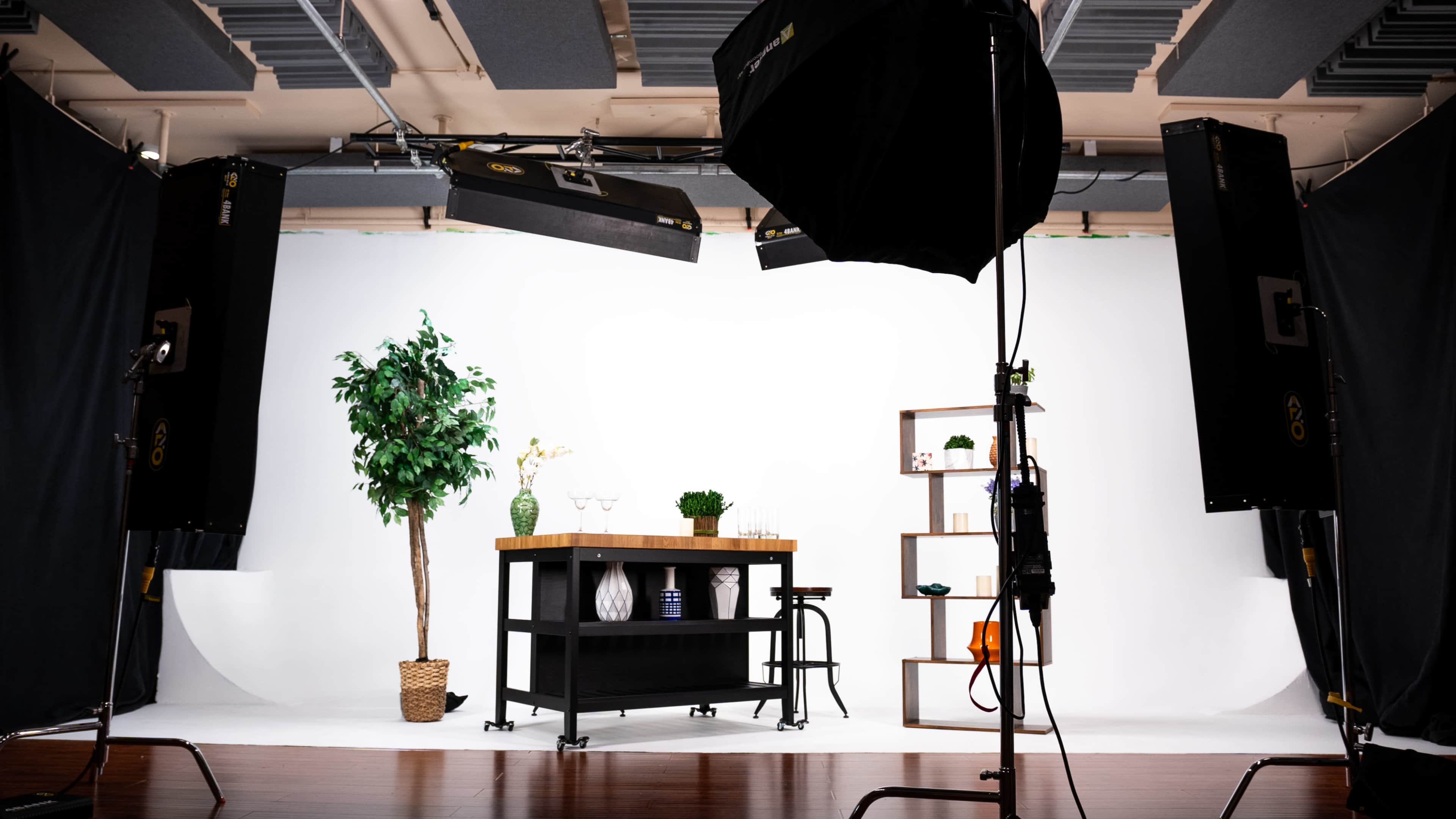 A photography studio setup with a work table, decorative plants, a shelf with various items, and lighting equipment arranged in front of a plain white backdrop.