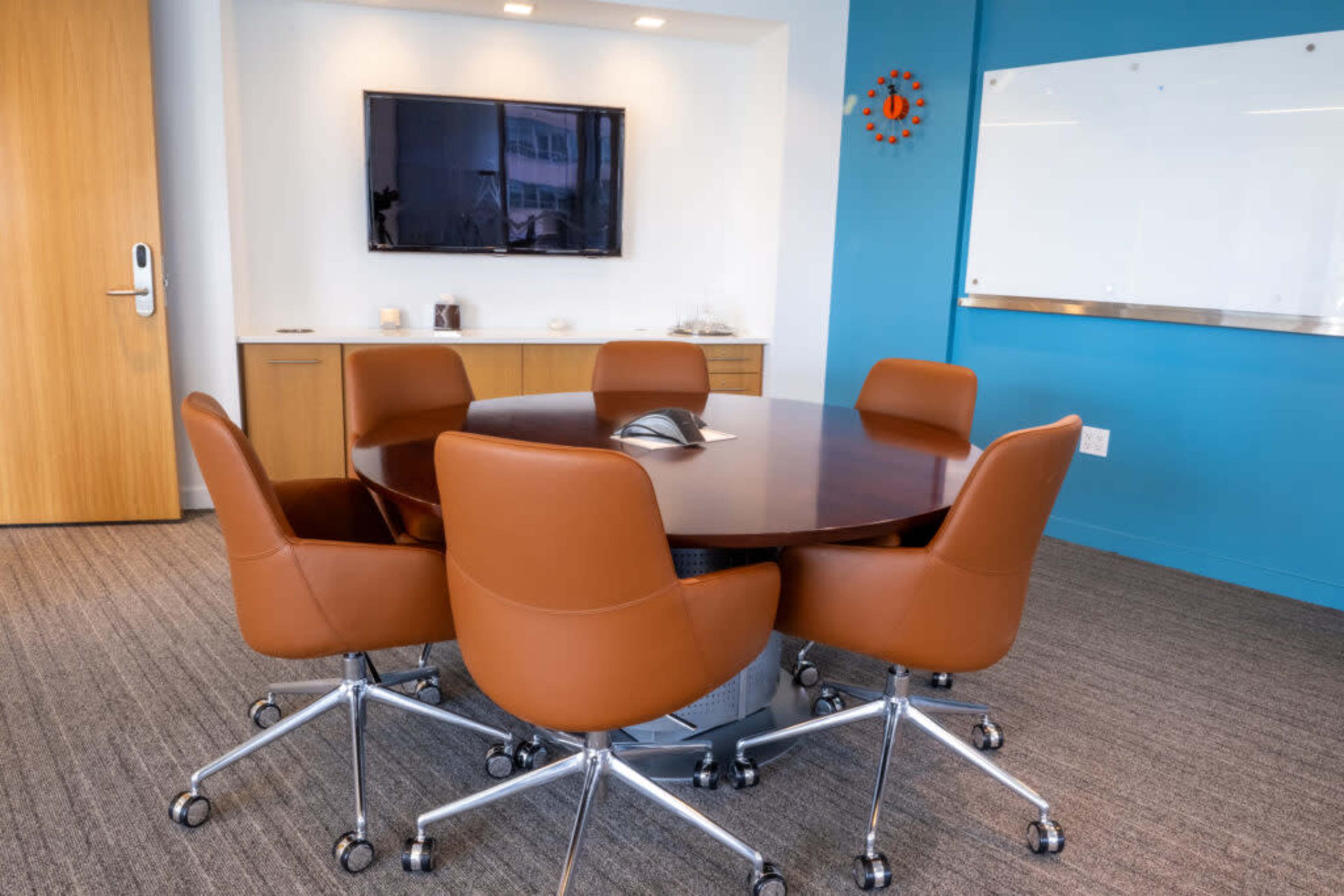 A modern conference room features a round wooden table surrounded by brown leather chairs and a wall-mounted television.