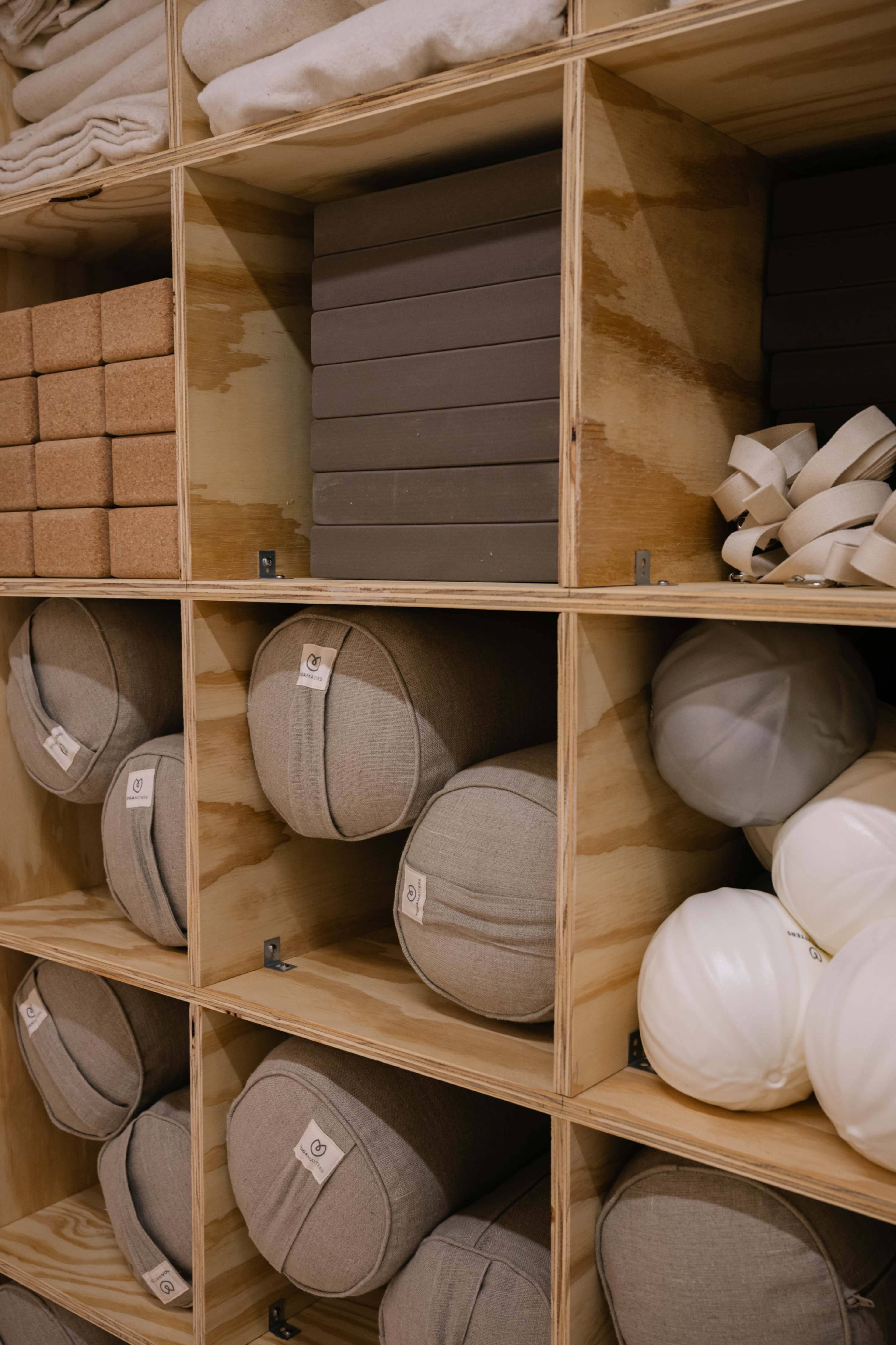The image shows a wooden shelving unit divided into square compartments containing various rolled mats, blocks, and cushions in neutral colors.