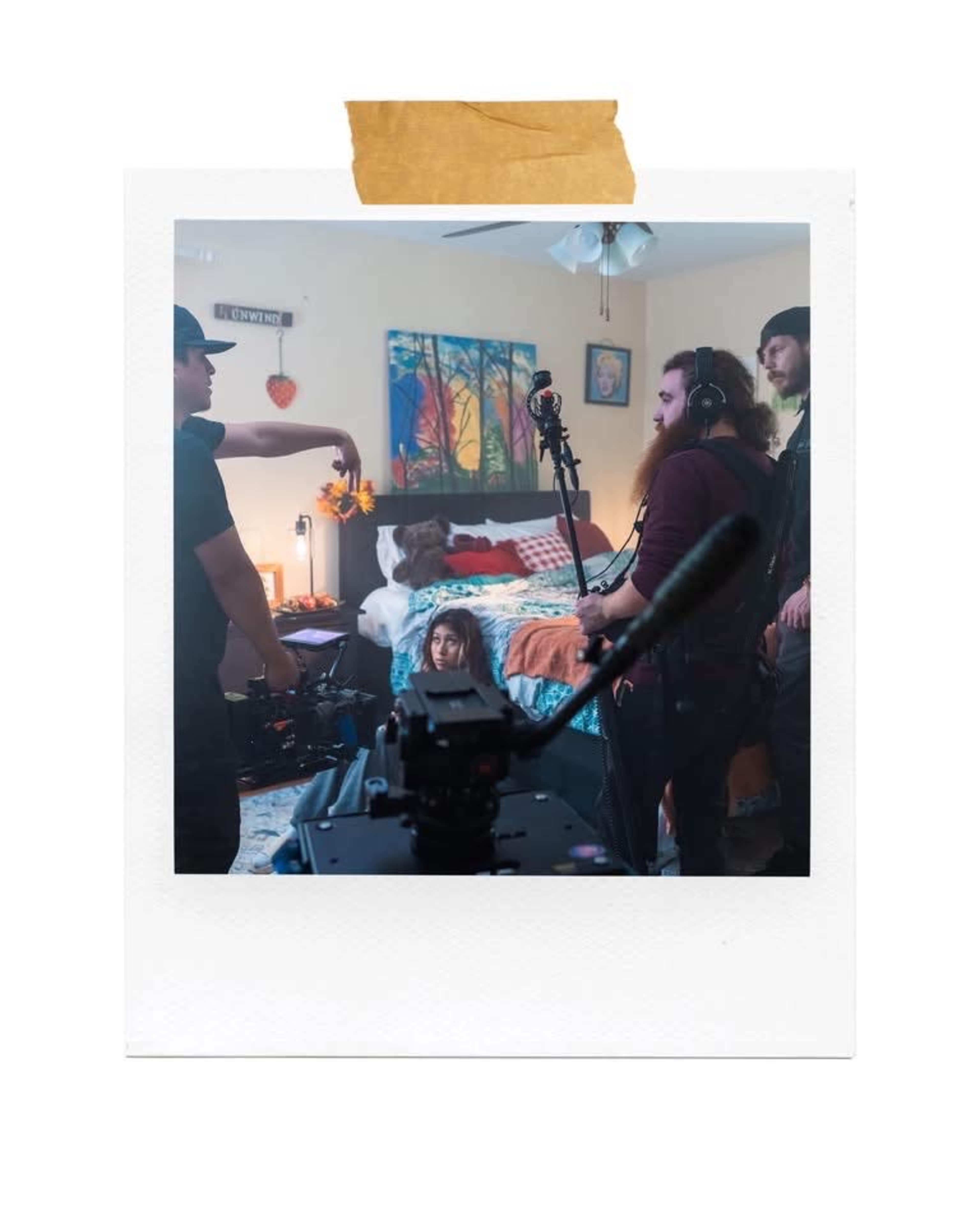 A film crew is set up in a bedroom, with one person directing while others prepare equipment and monitor the scene.