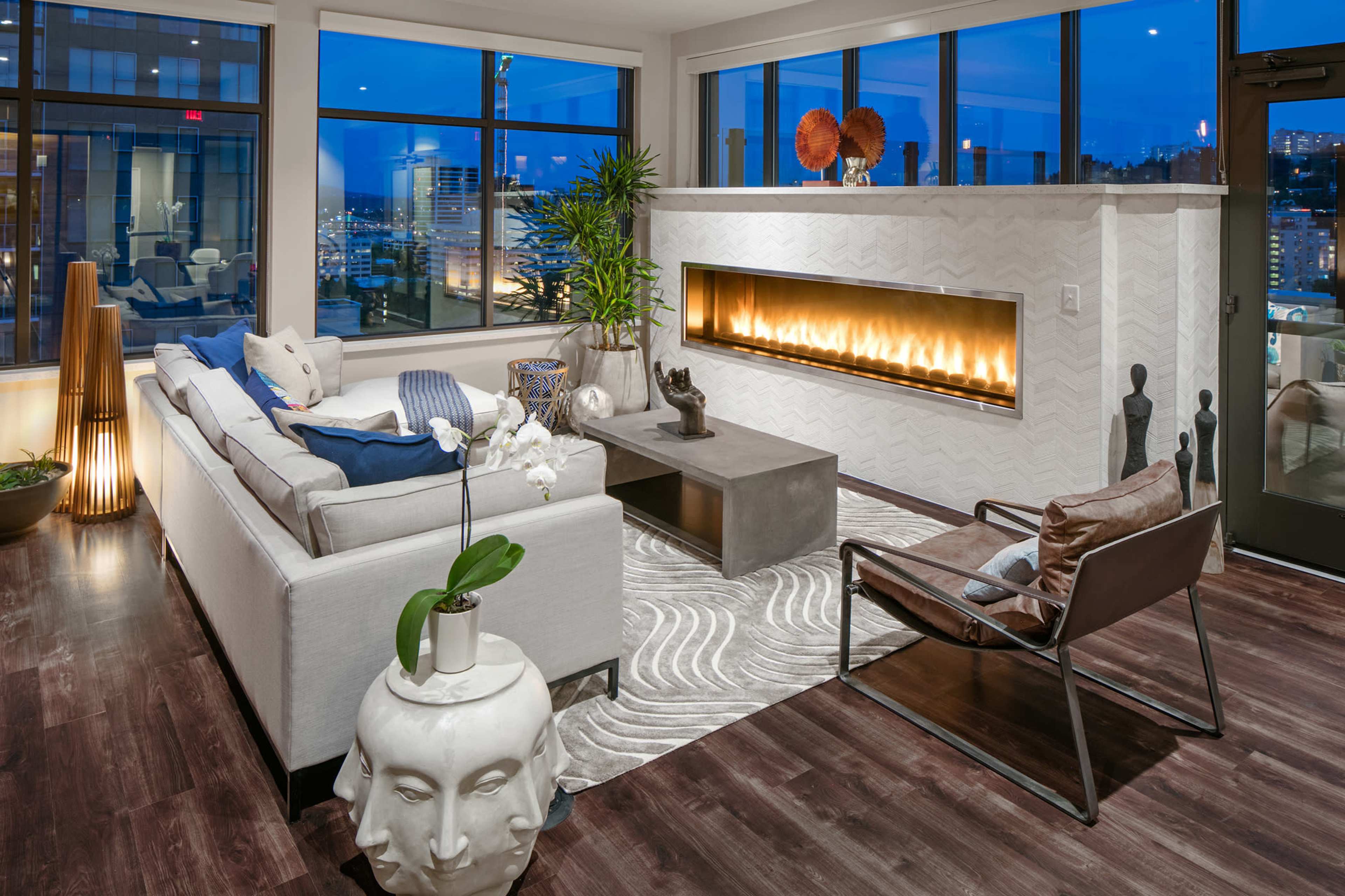 A modern living room features a sleek fireplace, large windows with city views, and stylish furniture arranged on a patterned rug.