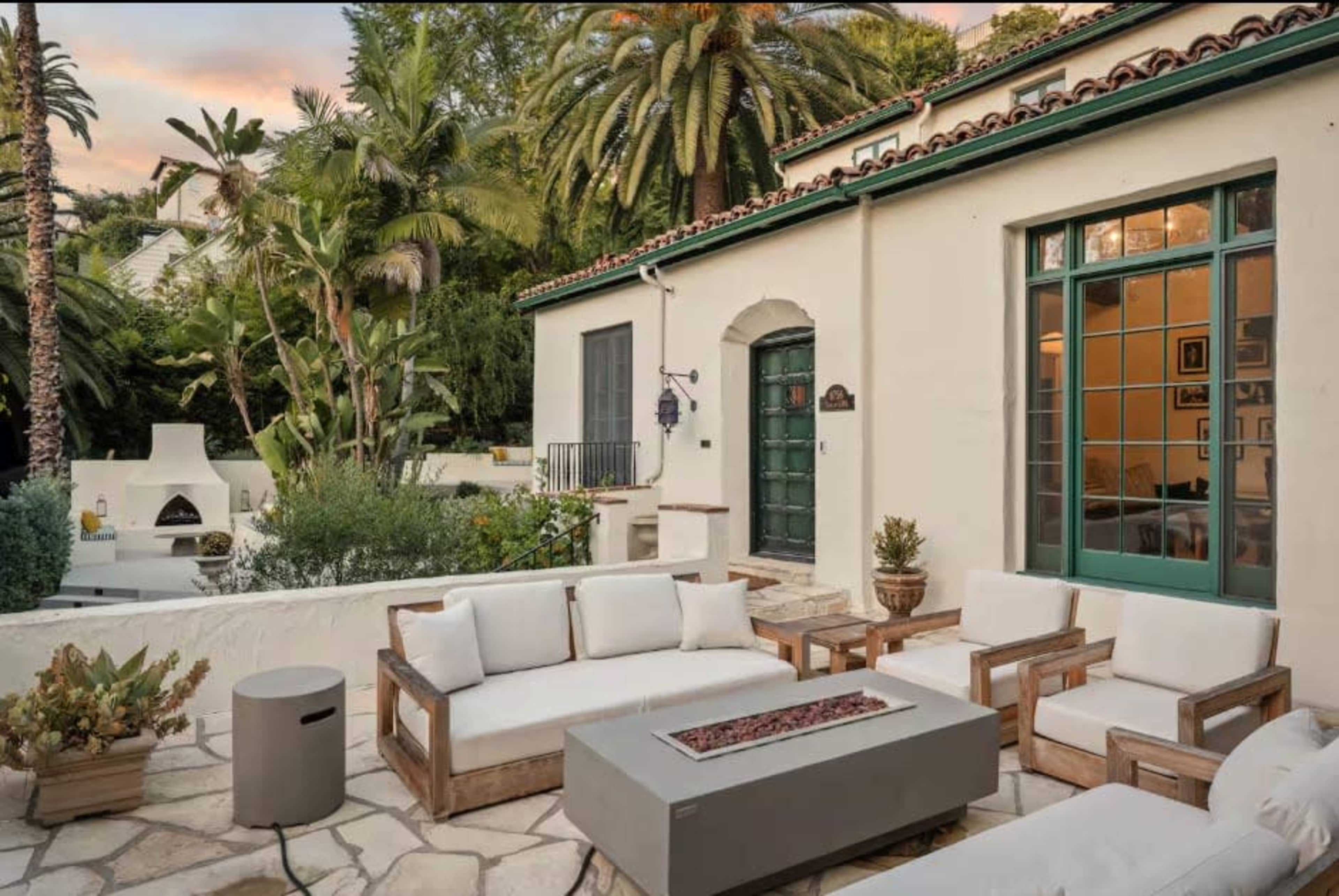 The image shows a patio area with a modern outdoor seating arrangement and a fire pit, surrounded by lush greenery and a house featuring a green door and large windows.