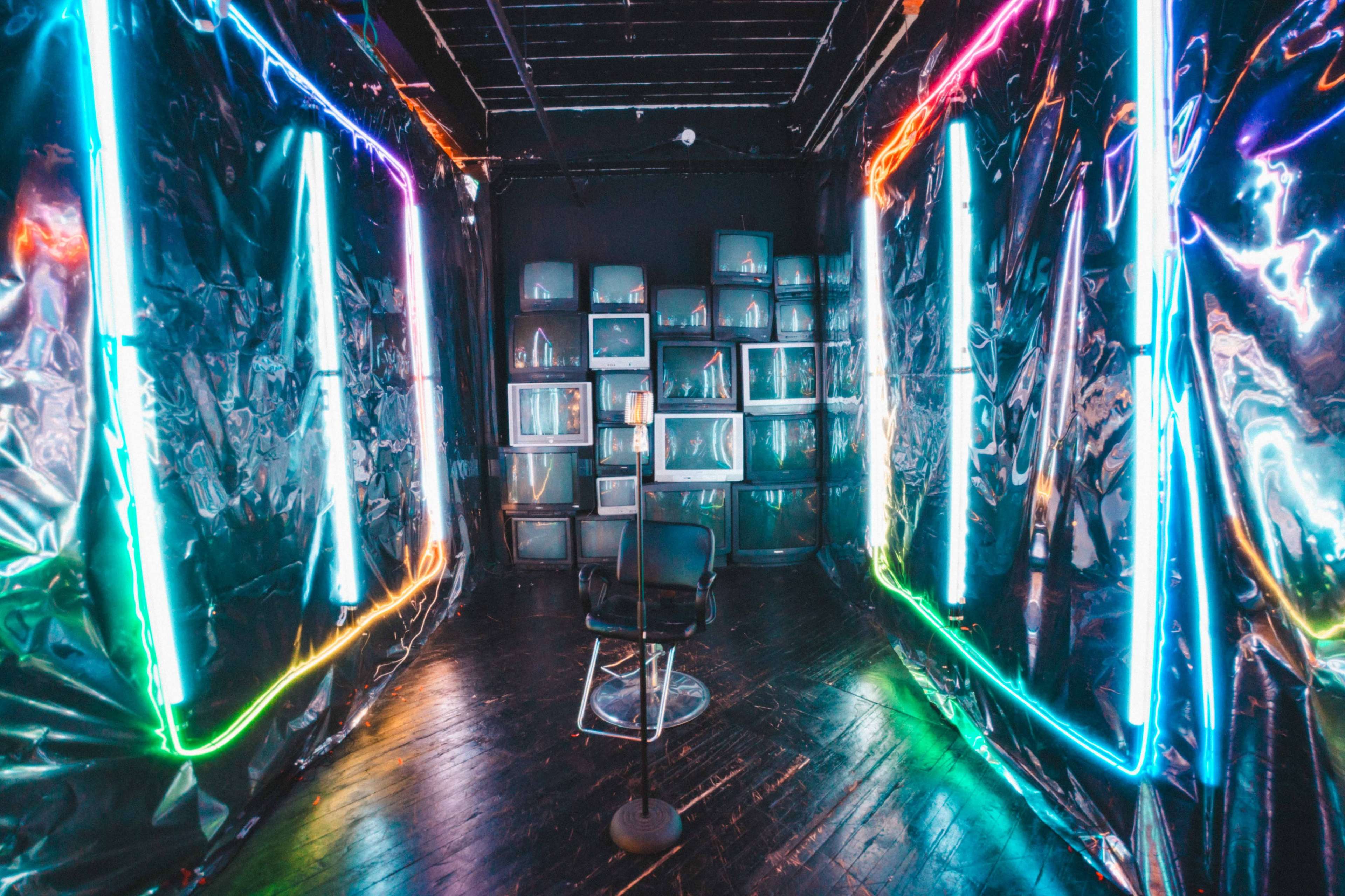 The image depicts a dimly lit room with walls covered in shiny black material, illuminated by colorful neon lights and featuring vintage television sets arranged on shelves.