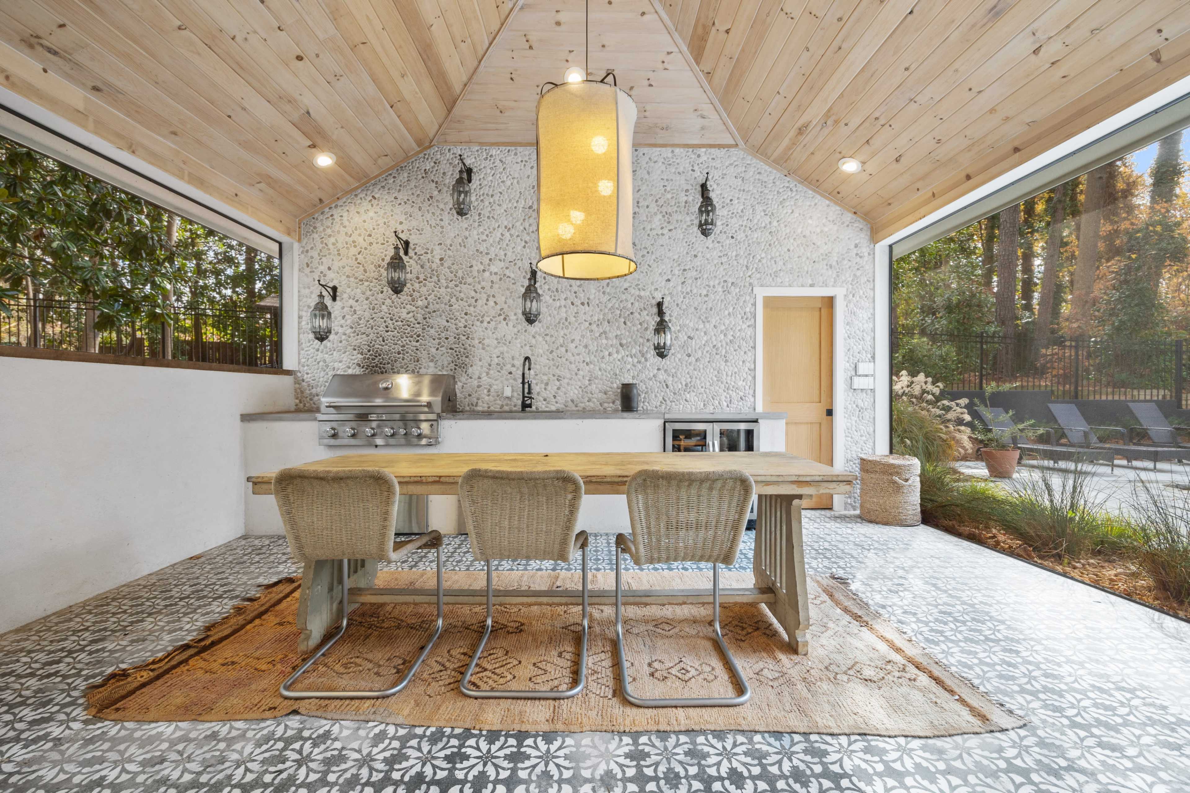 A modern outdoor kitchen with a wooden table, three wicker chairs, and a stone wall, surrounded by large windows and a patterned tile floor.