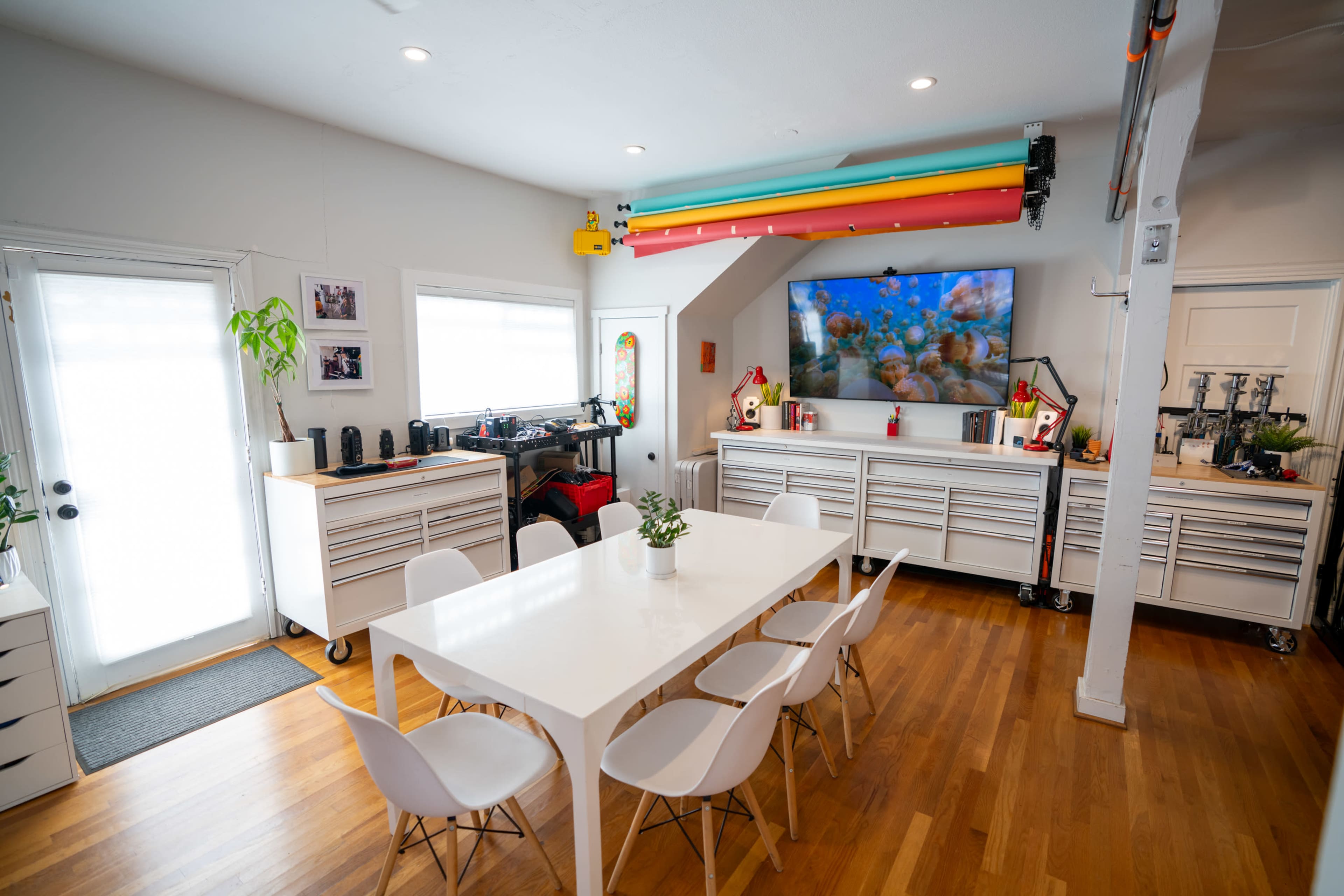 The image depicts a bright, organized workshop space featuring a large table surrounded by white chairs, workbenches with tools, and colorful overhead pipes.