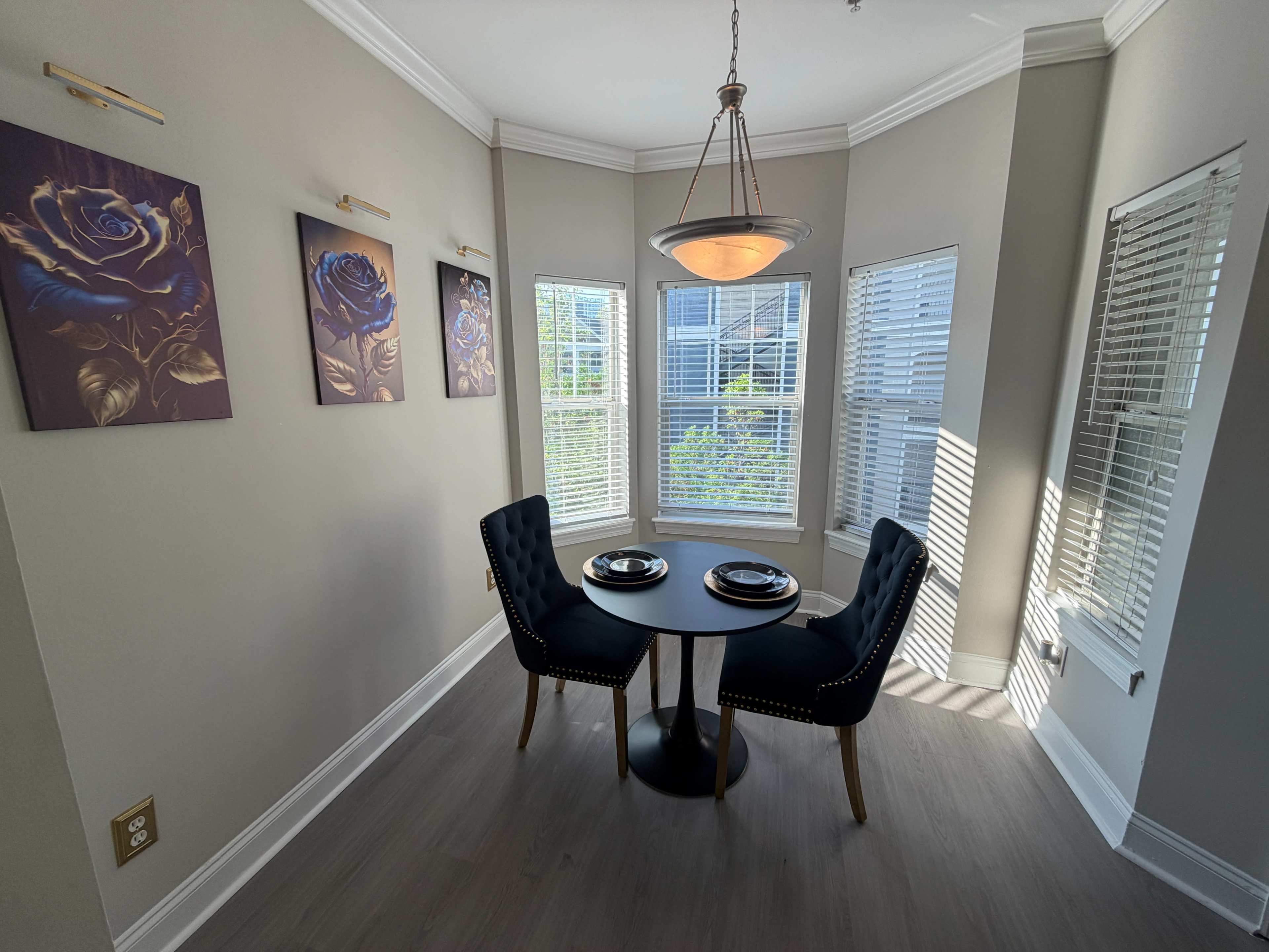 A small dining area features a round table with two upholstered chairs, surrounded by large windows and decorated with wall art of roses.