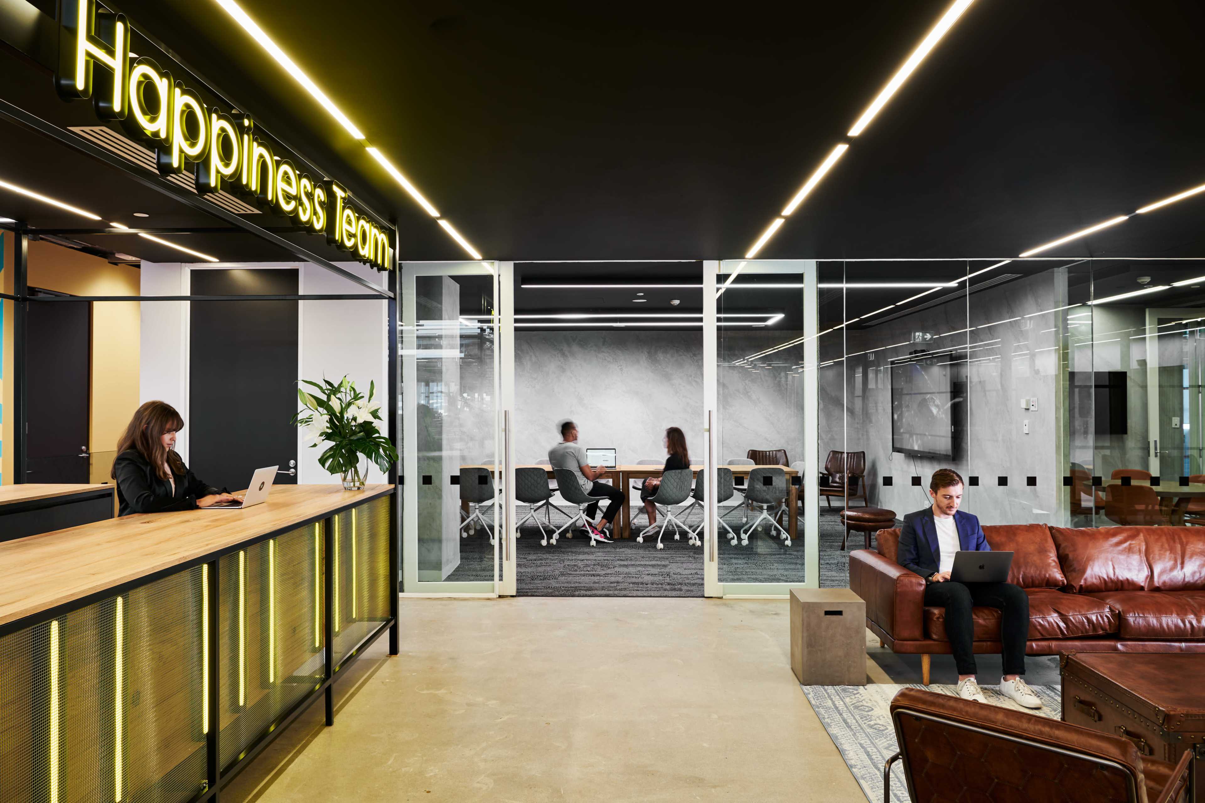 A modern office space features a reception area with "Happiness Team" signage, while a meeting room is visible in the background where people are collaborating.