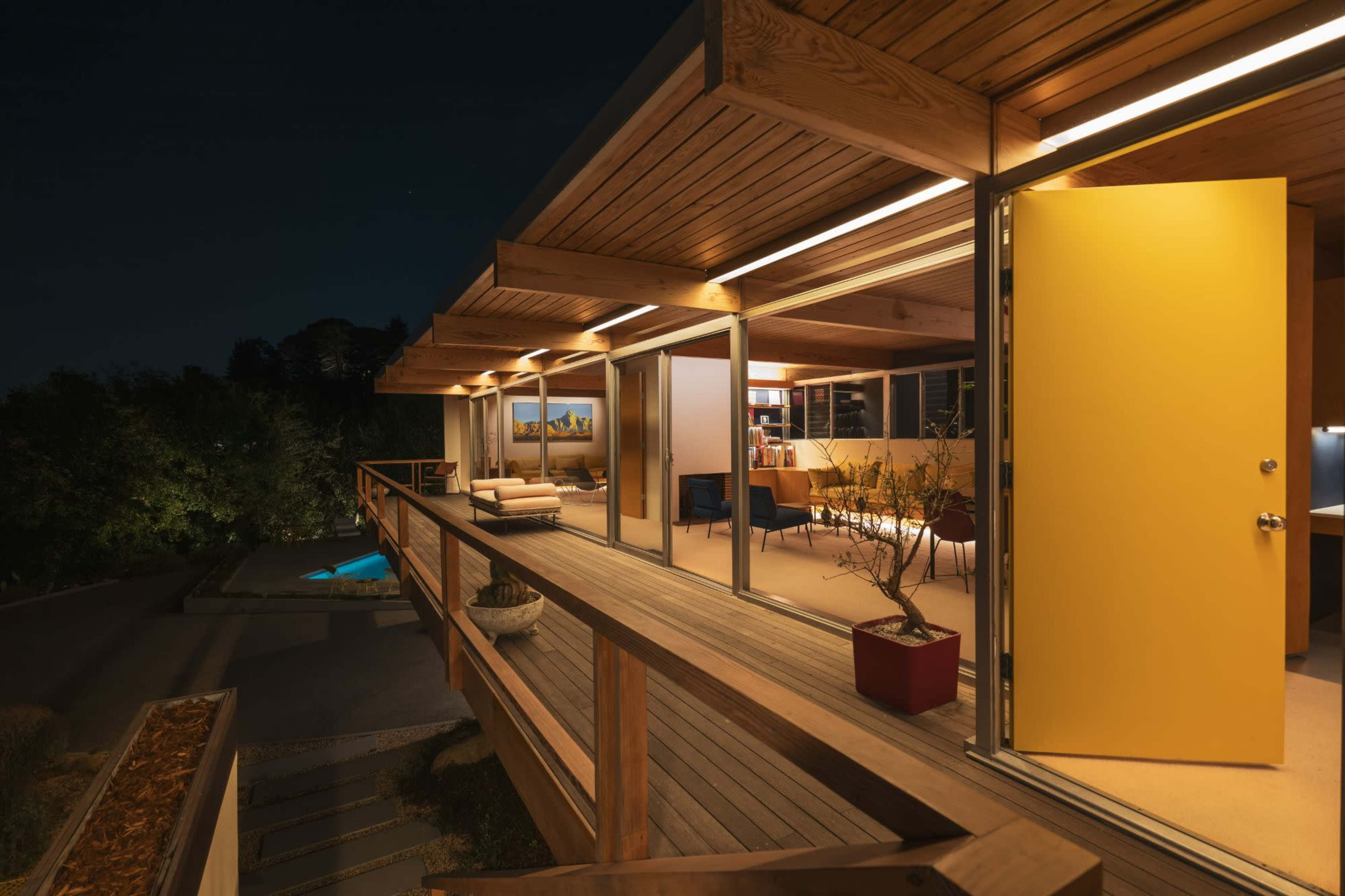 A modern wooden deck with a bright yellow door leads into a well-lit interior of a house at night, surrounded by greenery and a swimming pool.