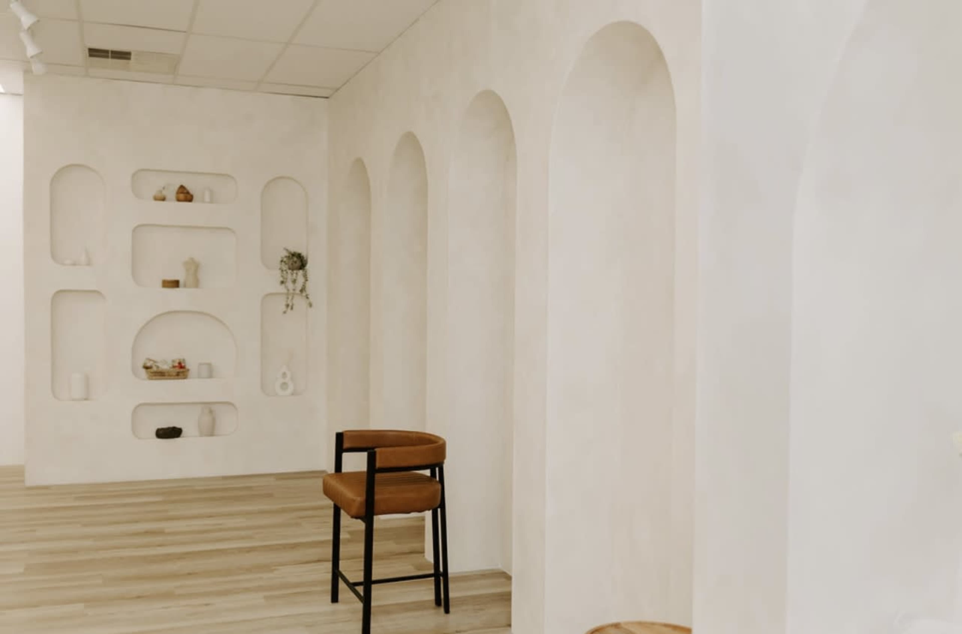 The image shows a minimalist, light-colored room with an arched wall display and a wooden stool.