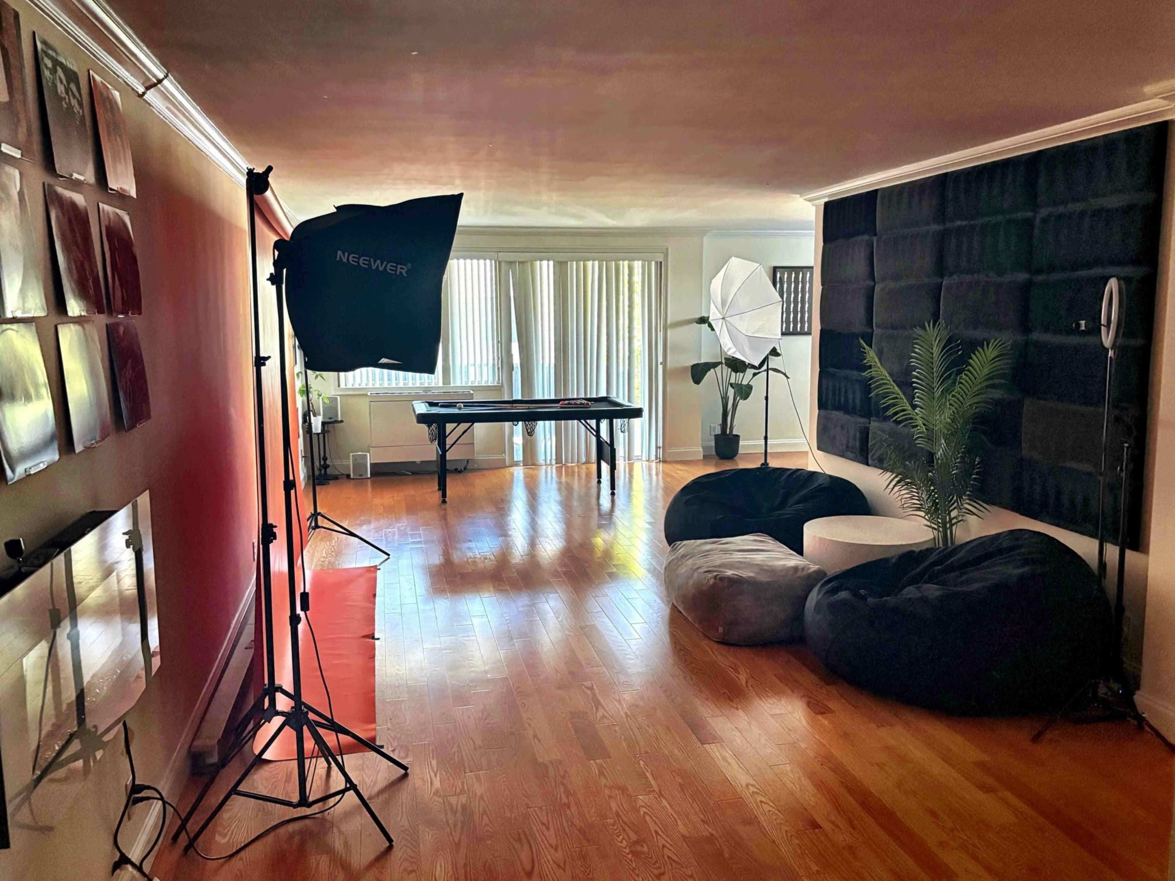 The room features a wooden floor with a large table, soft bean bags, photography lighting equipment, and a potted plant against a backdrop with textured walls.