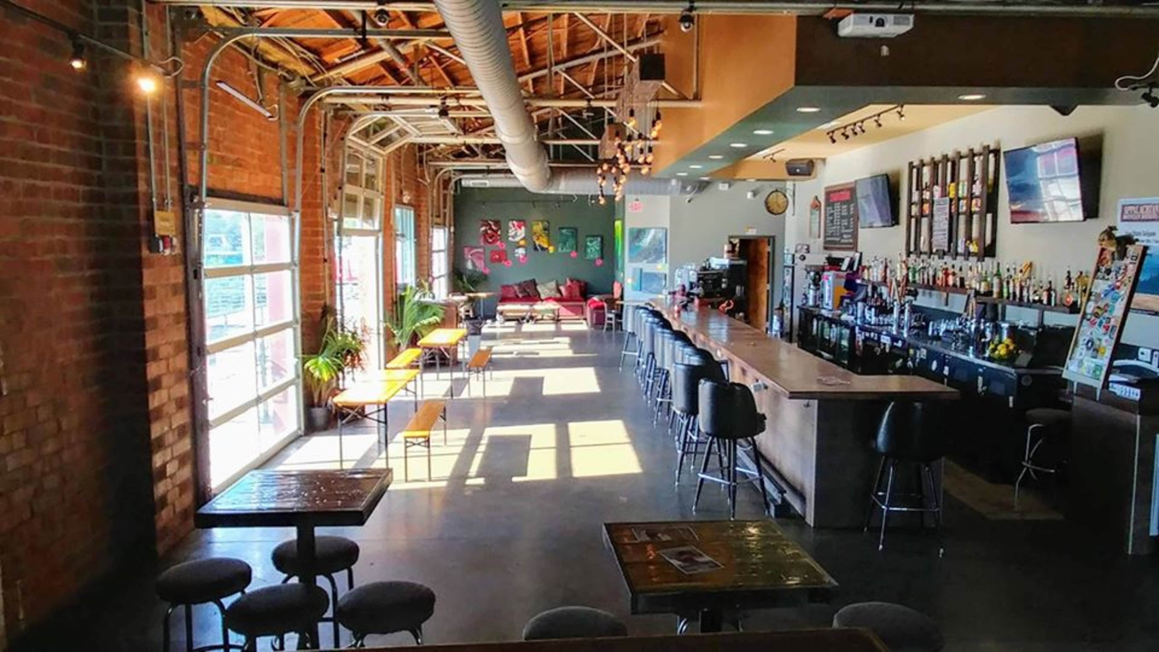 The image shows a spacious bar with a wooden counter, stools, and bright sunlight streaming through large windows, illuminating the interior featuring exposed brick walls and a mix of seating options.