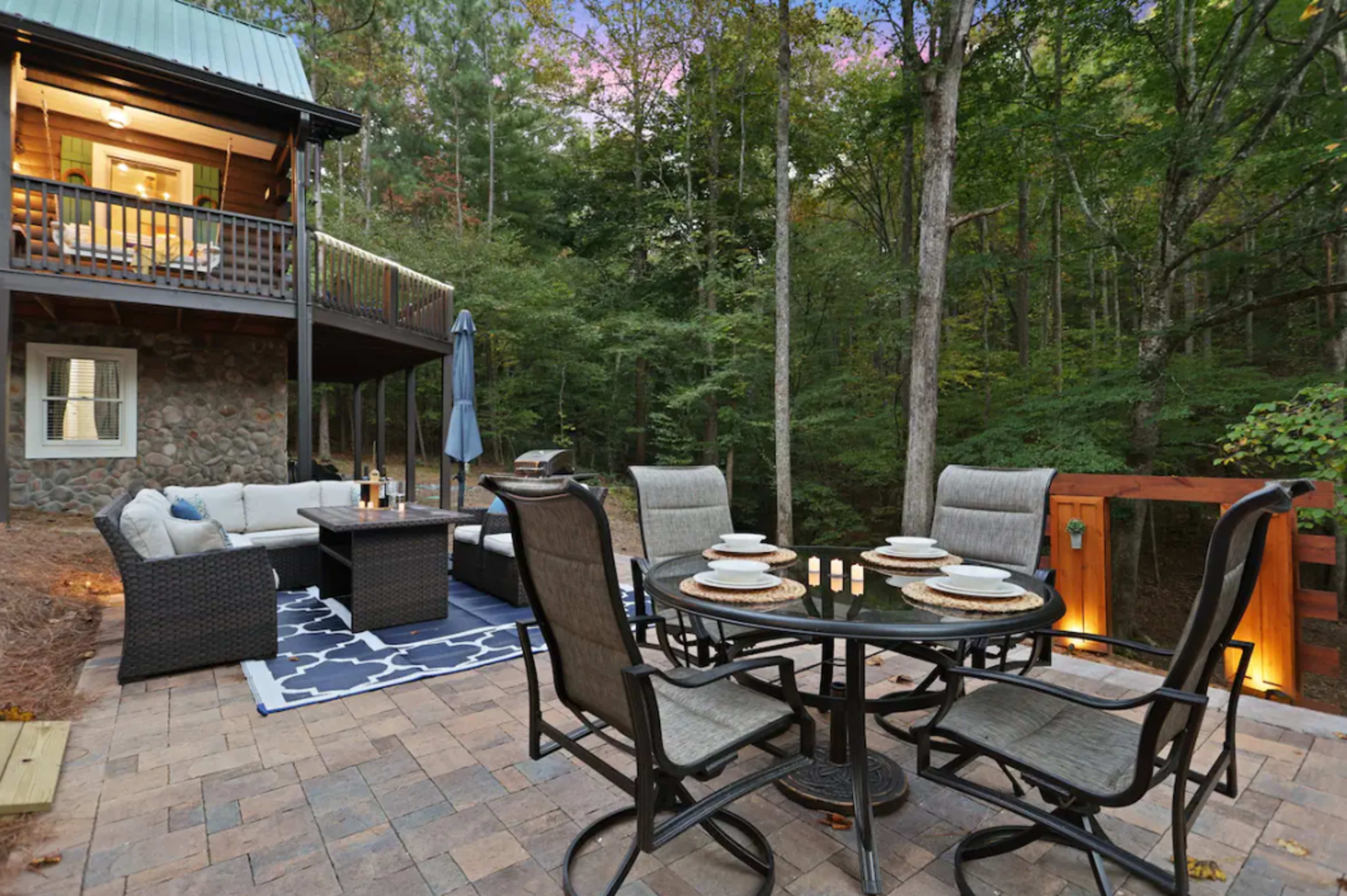 A patio with a dining table set for six chairs is surrounded by trees and features outdoor seating and a stone building in the background.