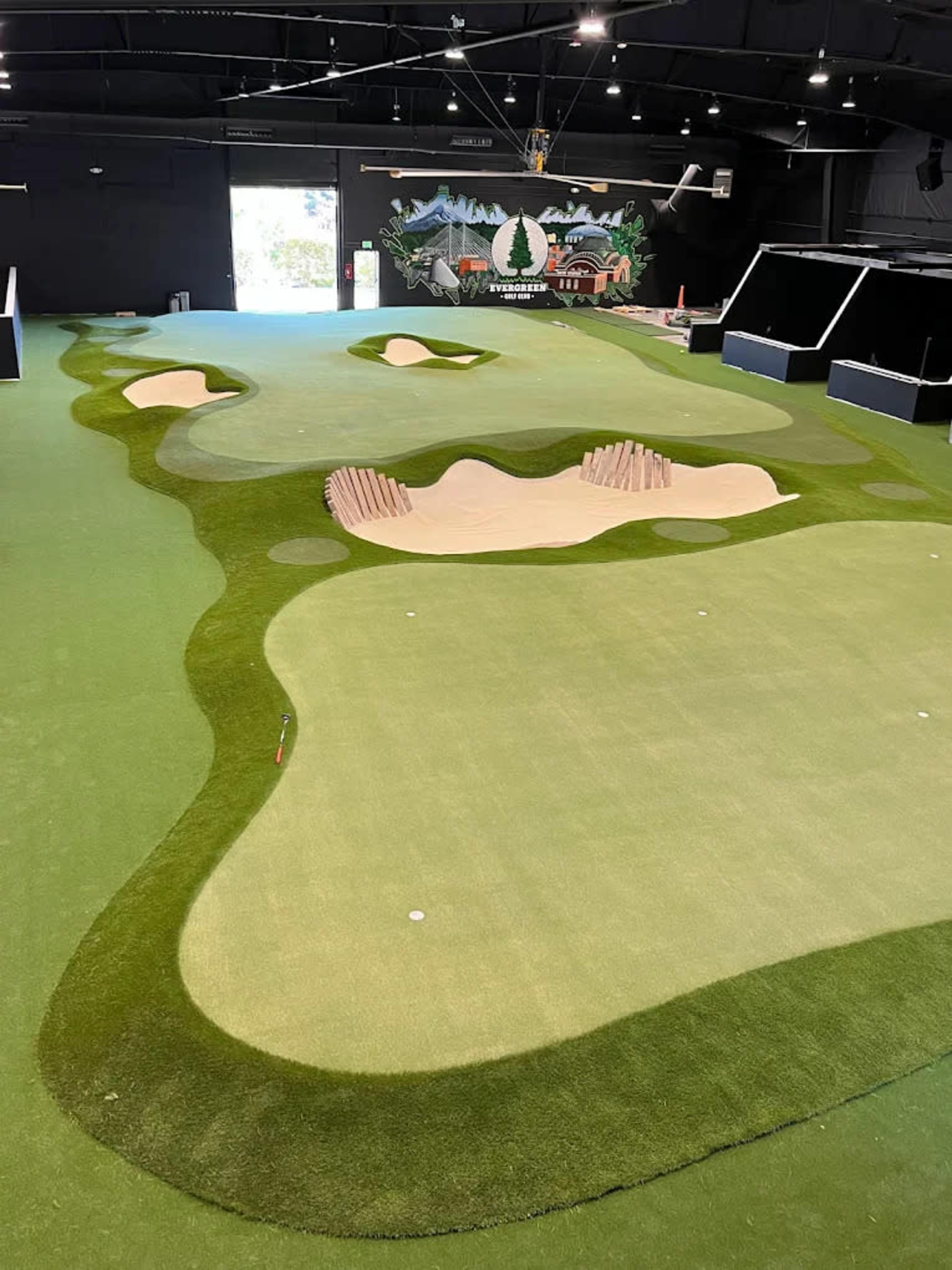The image shows an indoor golf facility with multiple putting greens and sand traps bordered by artificial turf.