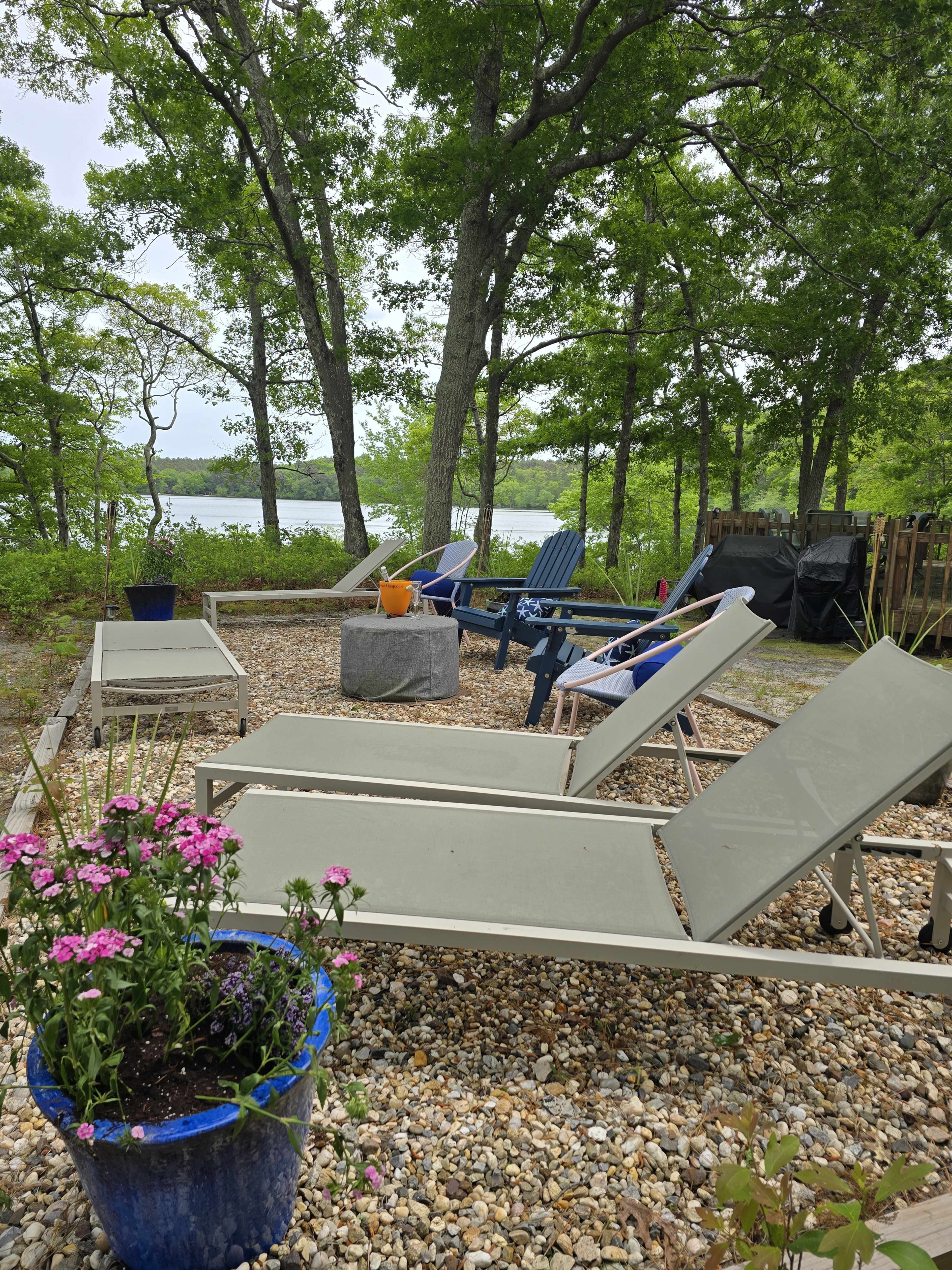 The image shows a patio area with multiple lounge chairs arranged around a circular table, bordered by trees and overlooking a body of water.
