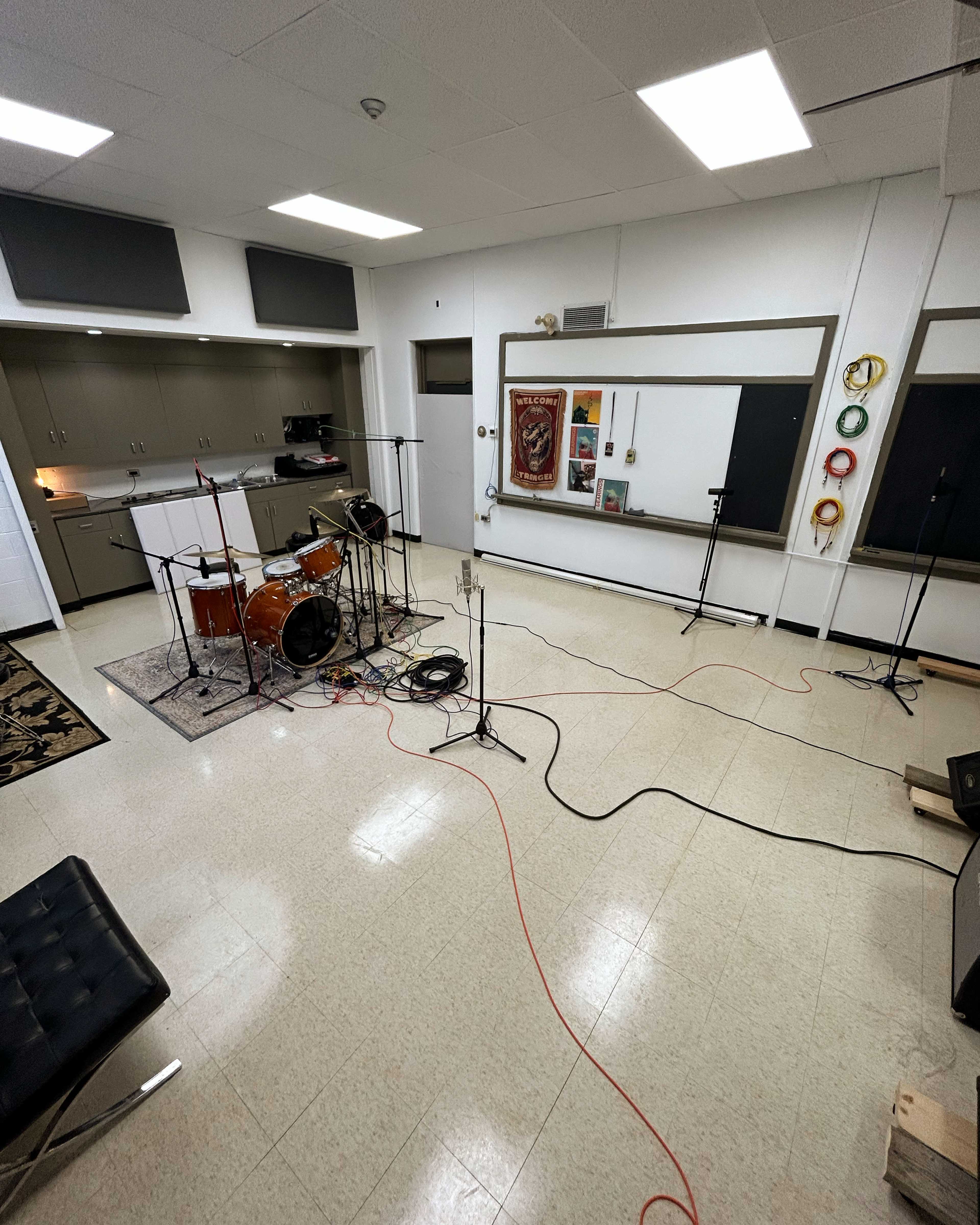 A room is set up for music recording, featuring a drum kit on a rug, multiple microphones, and cables scattered across the floor.