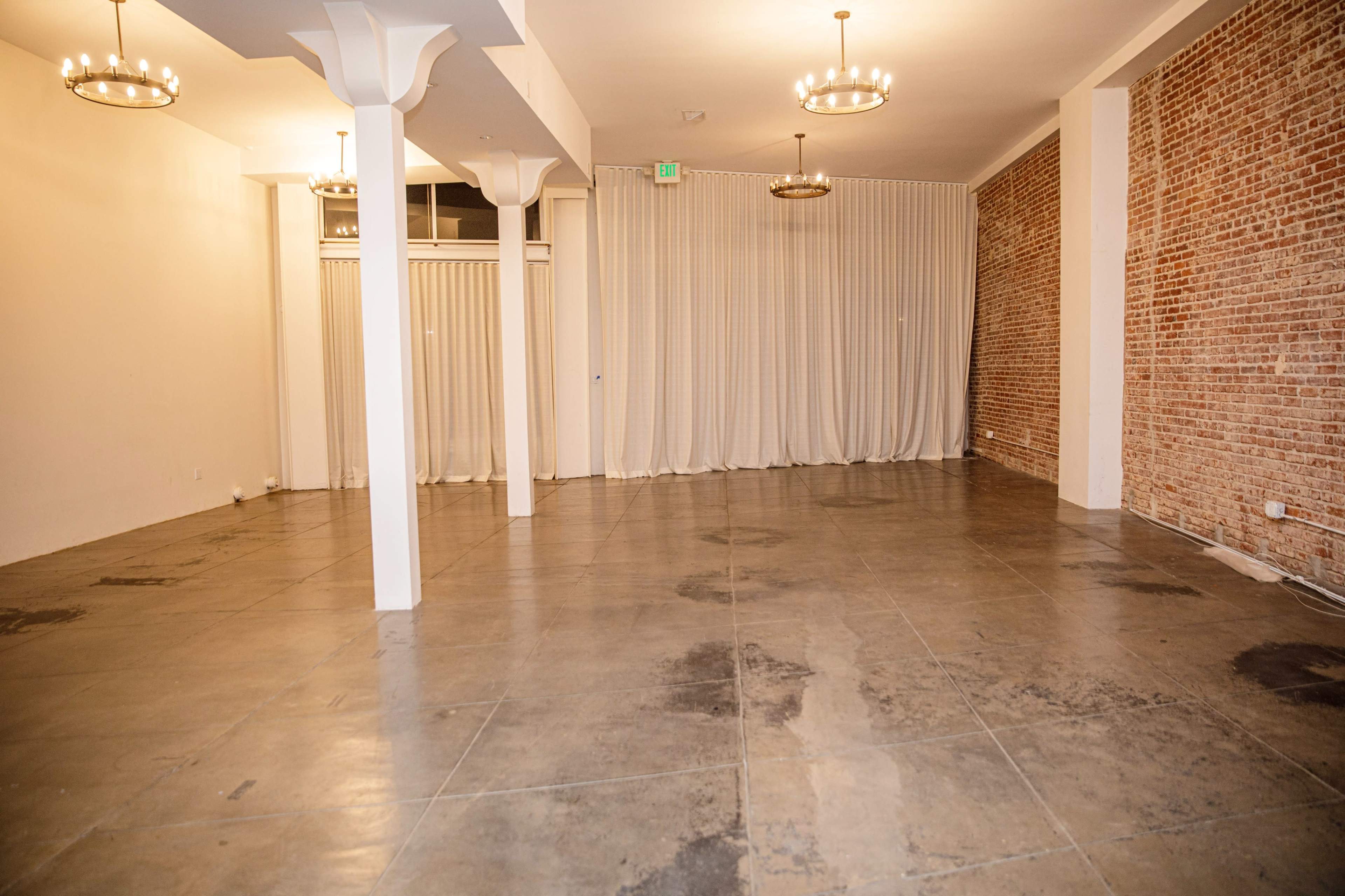 The image shows an empty room with concrete flooring, exposed brick walls, and hanging light fixtures.