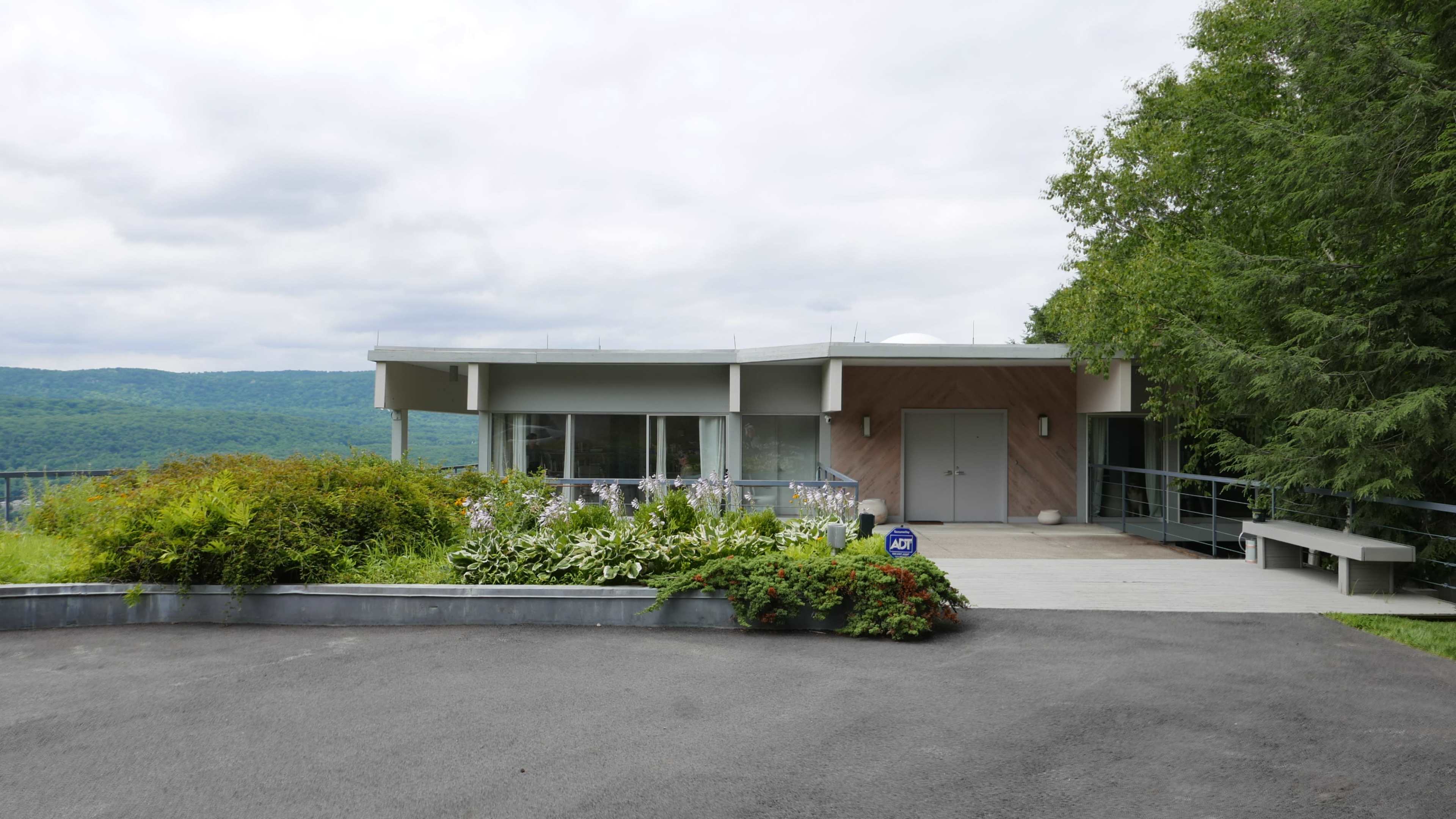 A modern single-story house with large windows is surrounded by greenery and positioned on a slight elevation overlooking a valley.