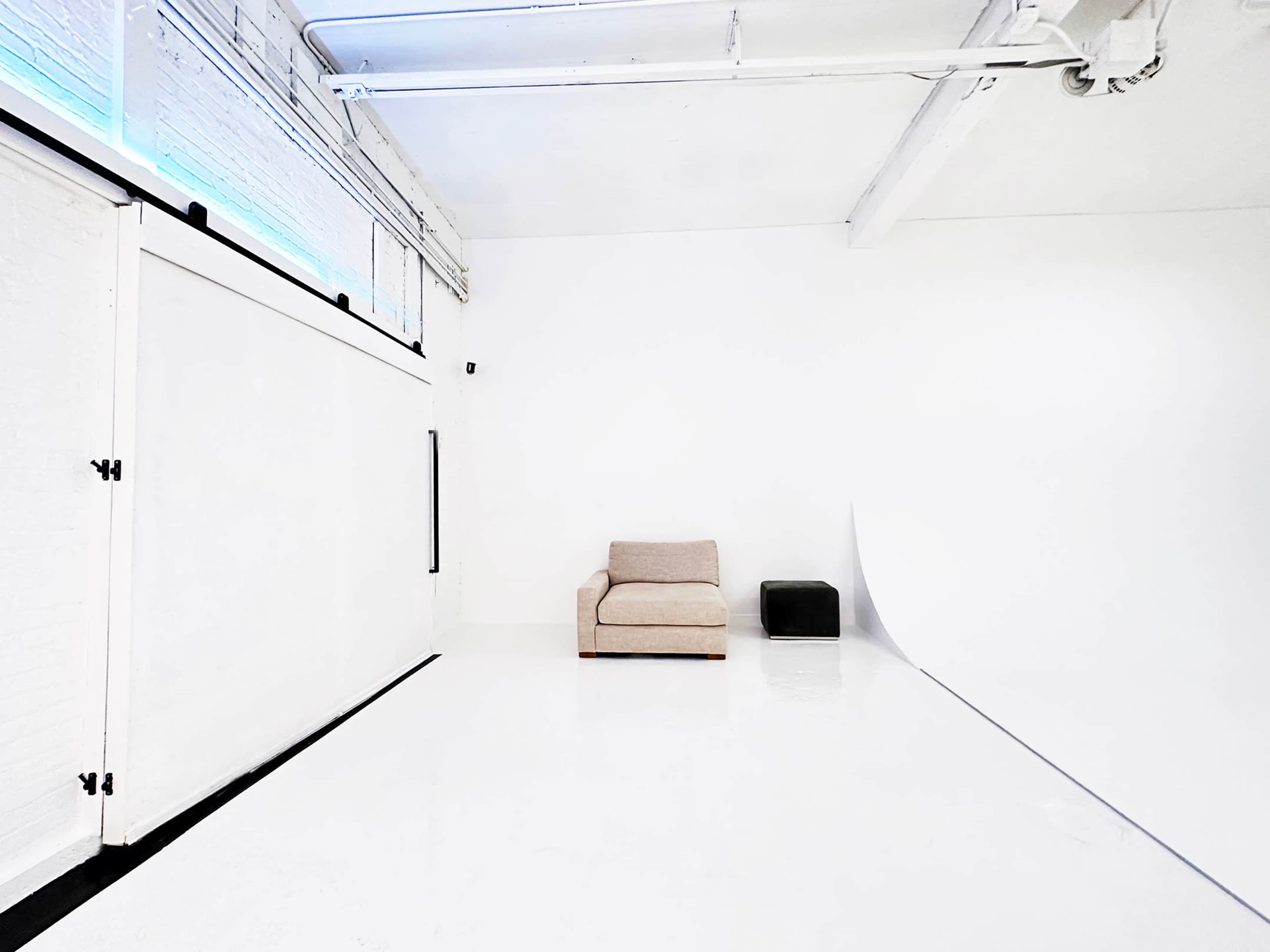 The scene features a minimalist studio space with a beige armchair and a small black stool against a backdrop of white walls and floor.