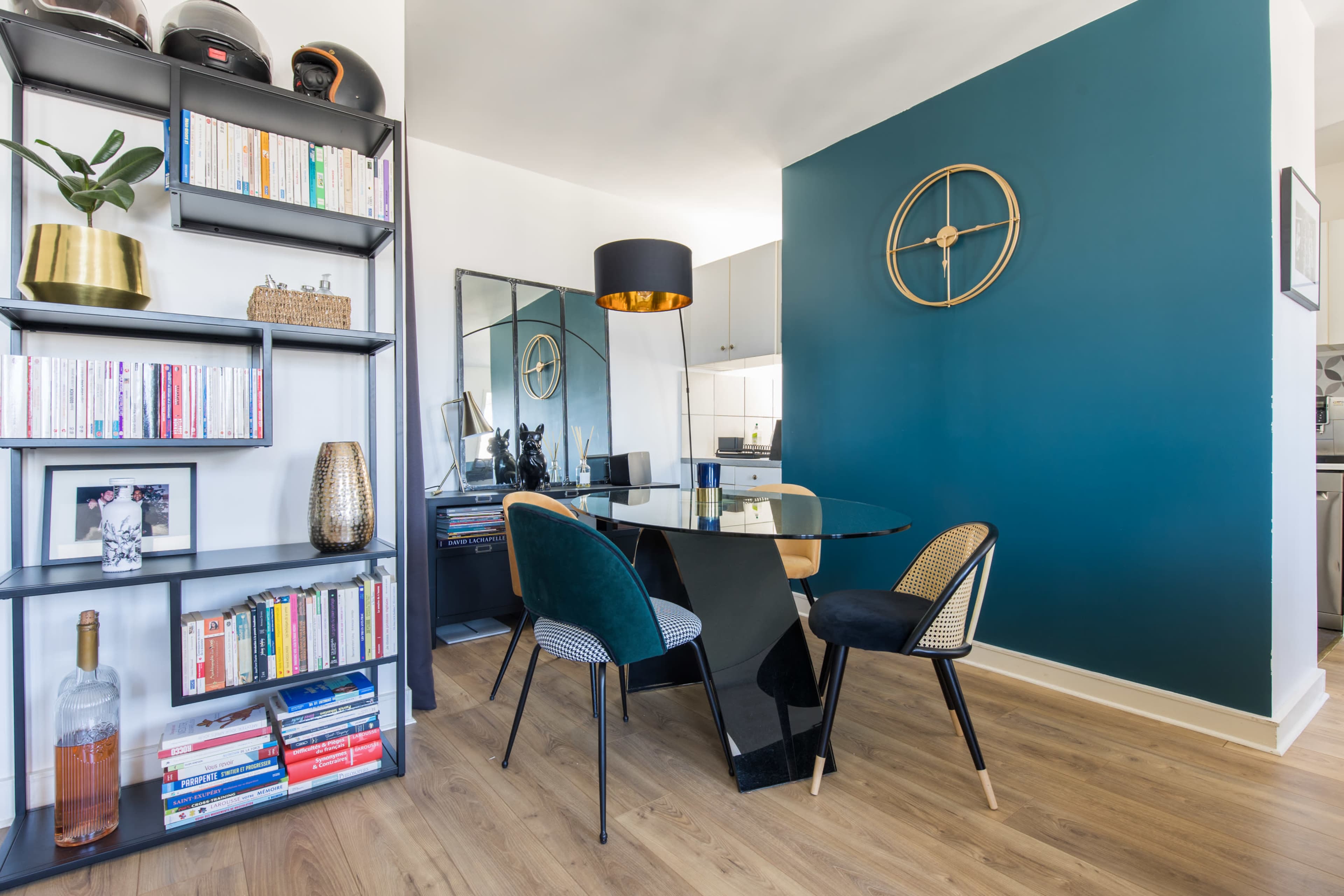 A modern dining area features a glass table surrounded by four chairs, with a dark blue accent wall and a bookshelf filled with books and decorative items.