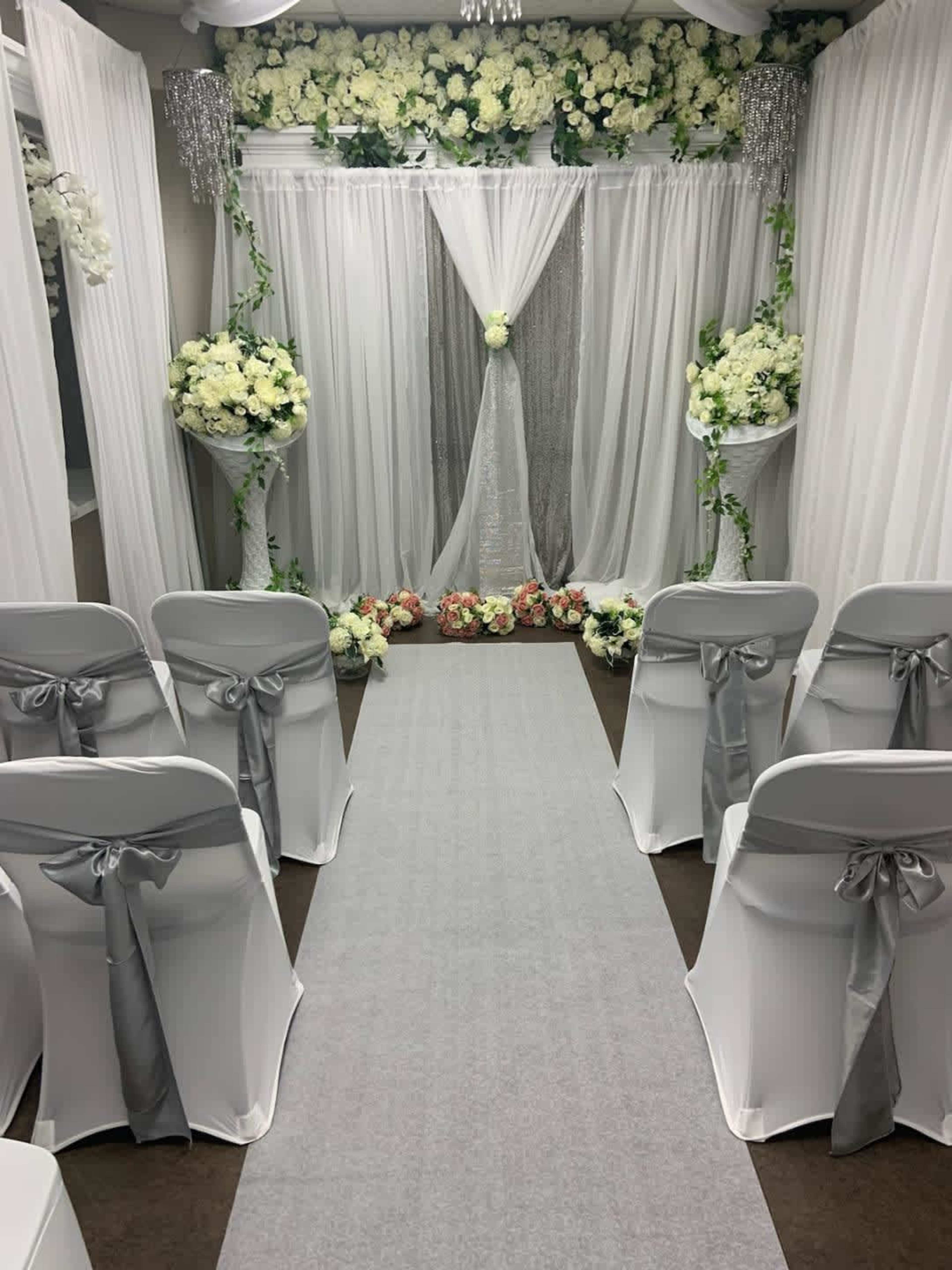A wedding ceremony setup features rows of chairs adorned with silver ribbons facing a floral-decorated backdrop and a white draped altar.
