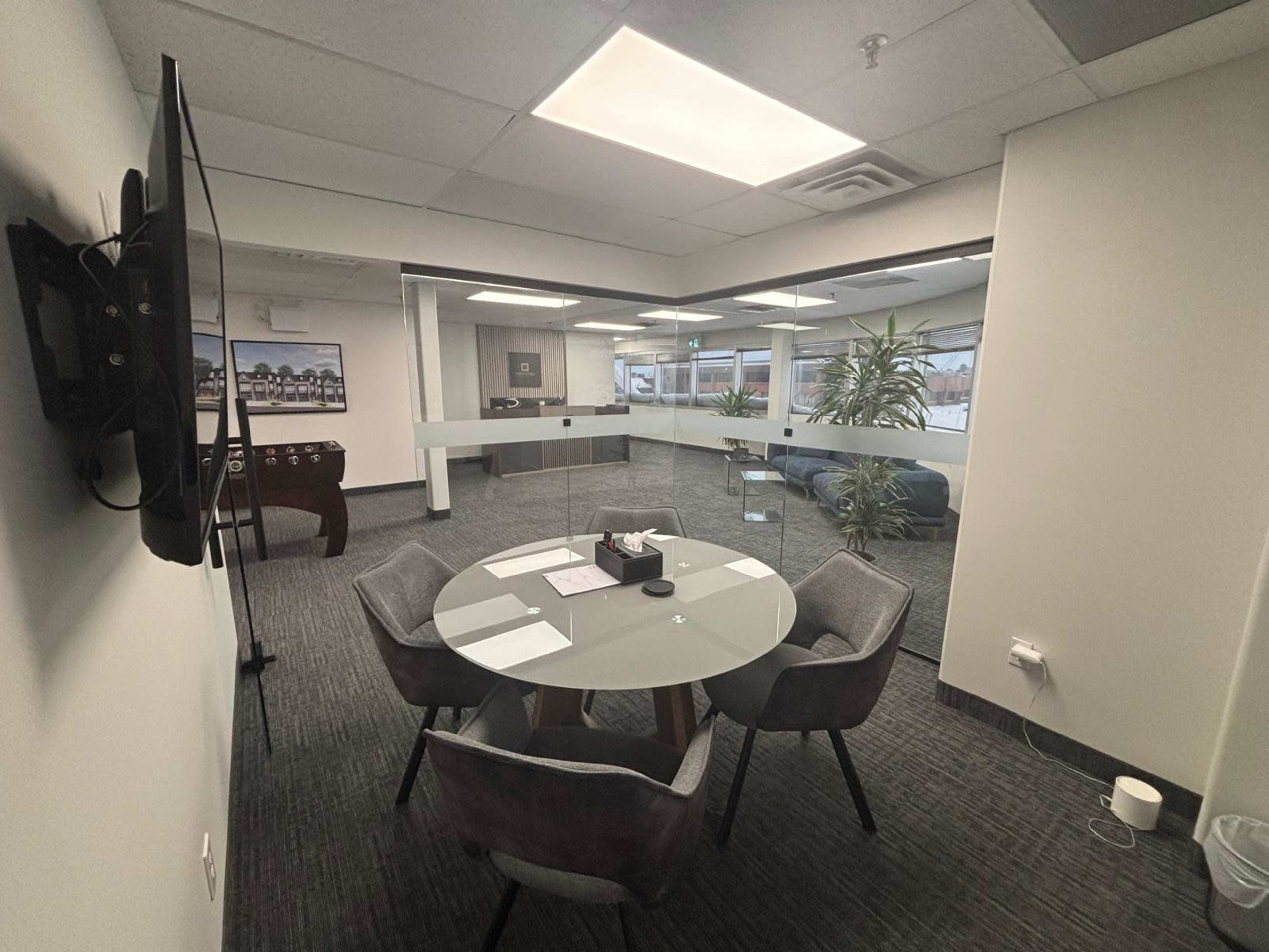 A modern meeting room features a glass table surrounded by four chairs, with a television mounted on the wall and large windows letting in natural light.