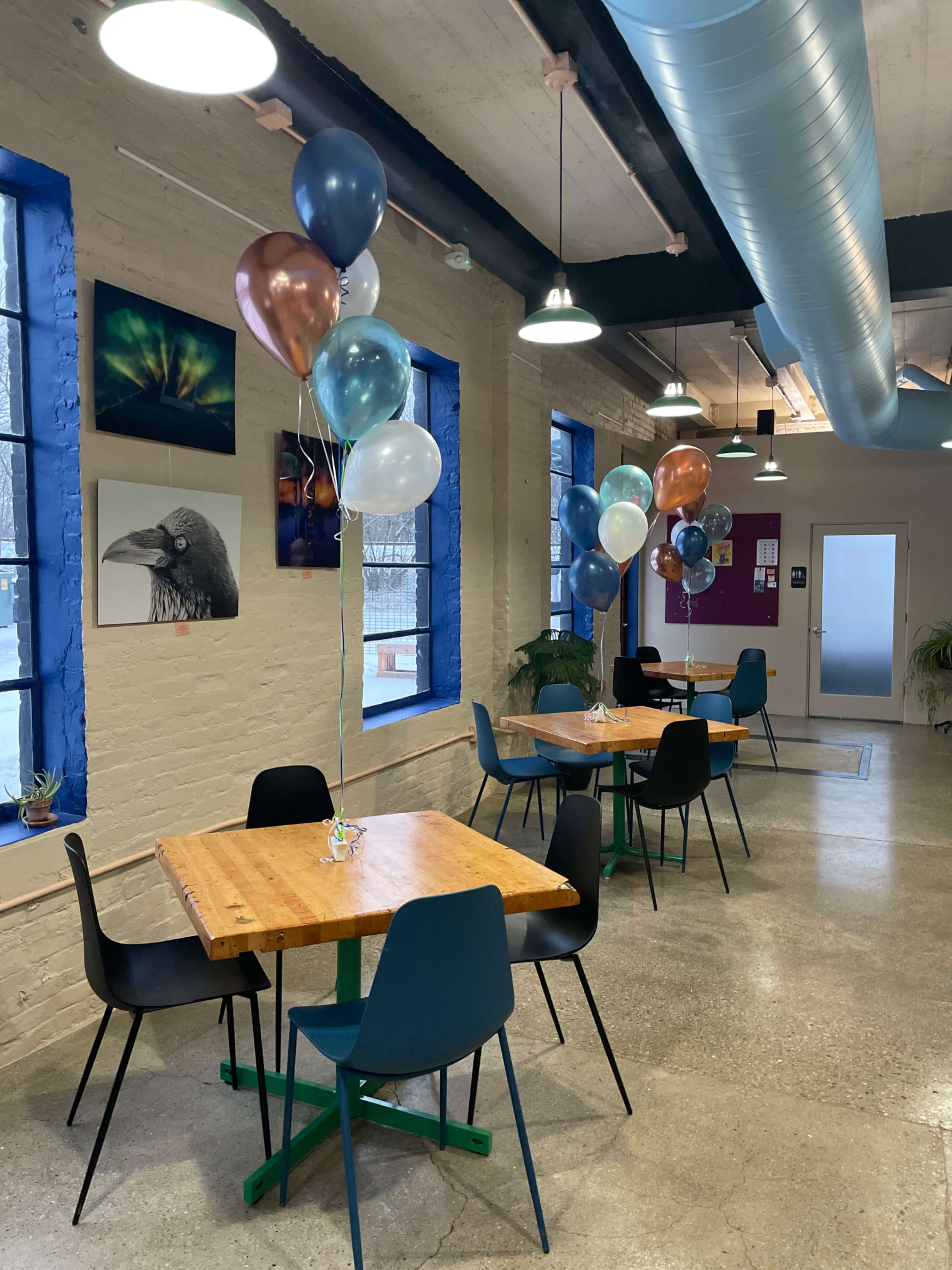 A modern café features several wooden tables surrounded by chairs and is decorated with clusters of balloons in various colors.