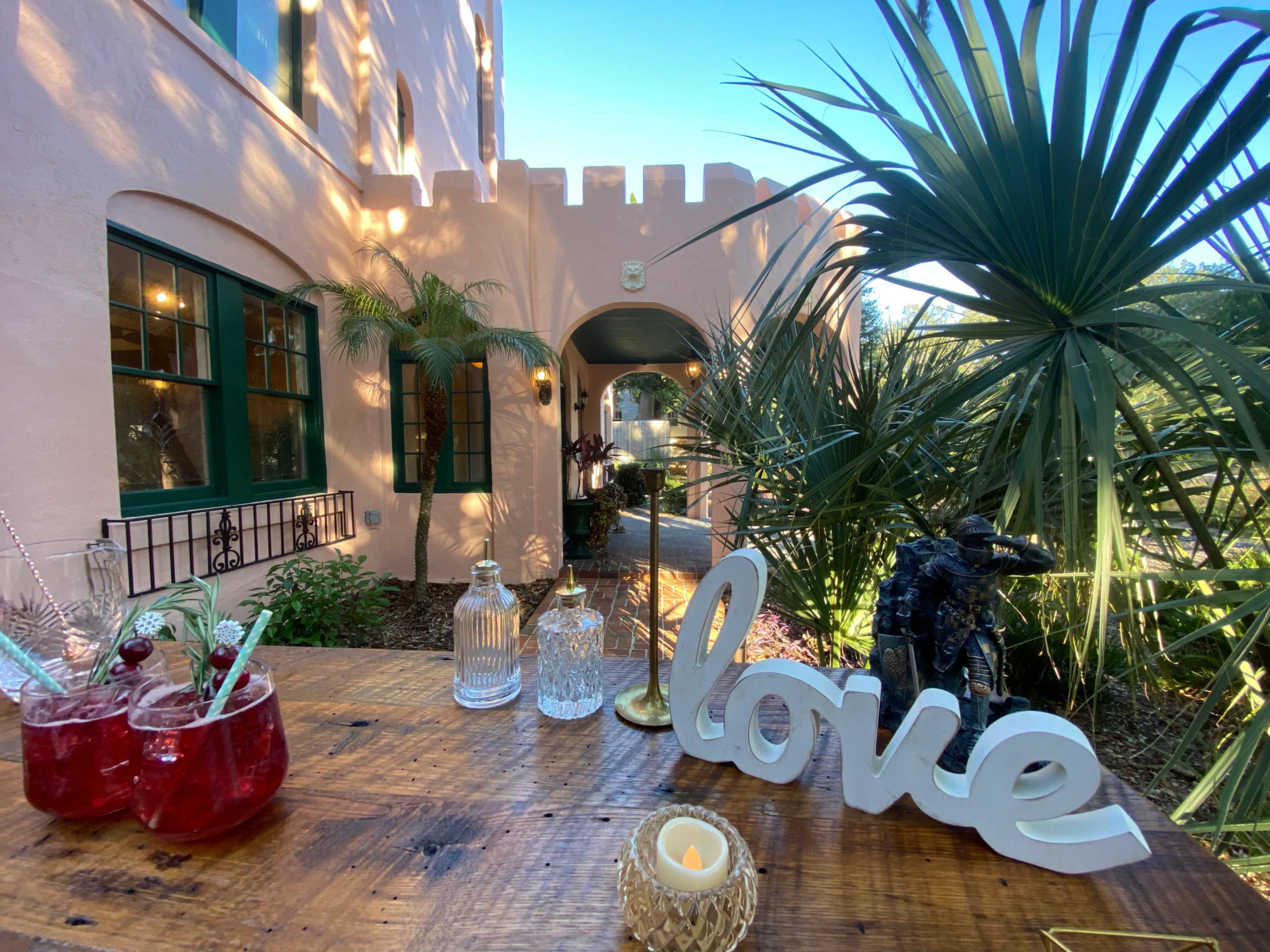 A wooden table with drinks and a decorative "love" sign overlooks a garden with palm plants and a castle-like building in the background.