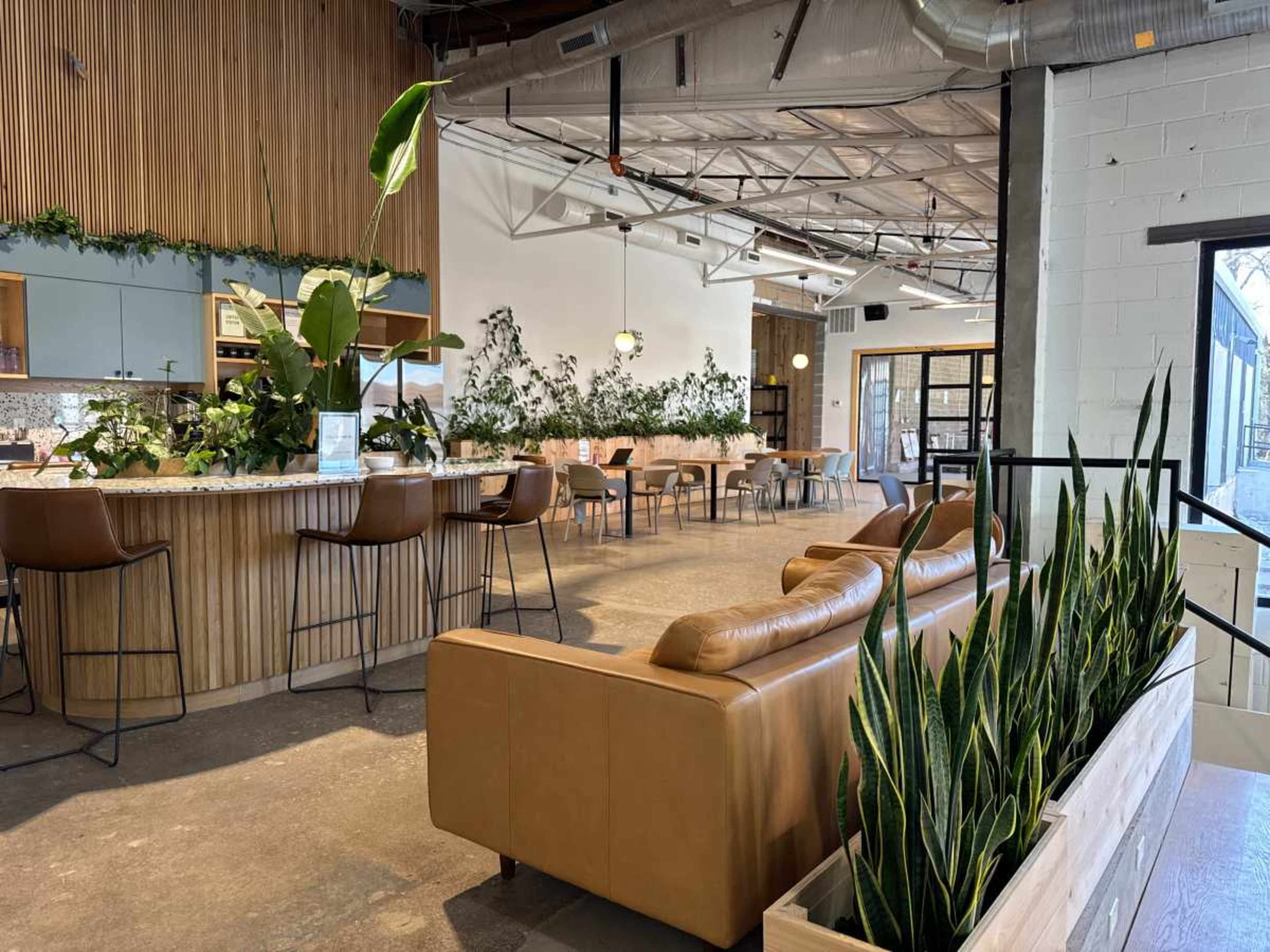 The image shows a modern interior space featuring a bar with seating, tables arranged throughout, and indoor plants integrated into the design.