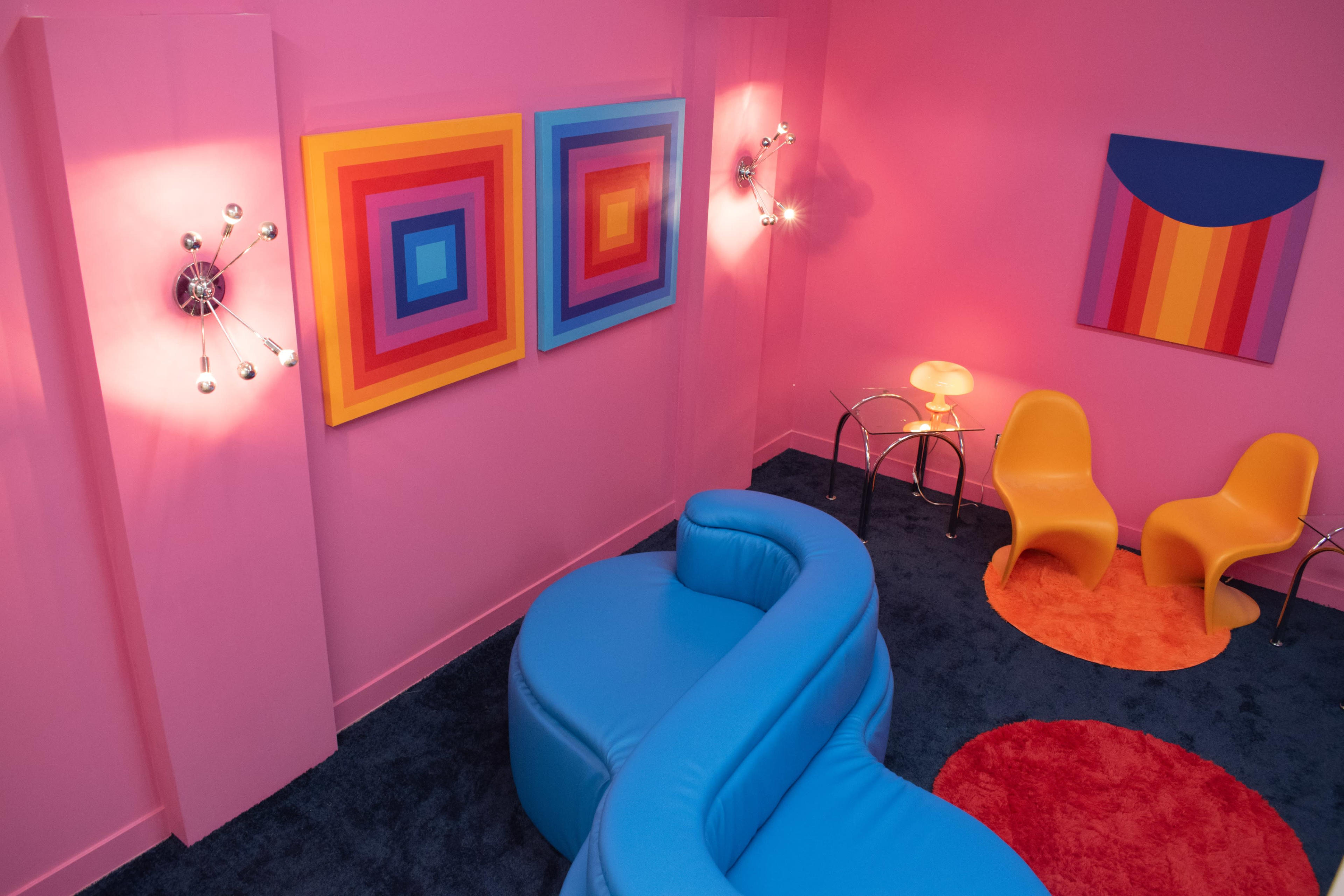 A colorful interior featuring a pink wall, a blue curved sofa, two orange chairs, and abstract artwork.
