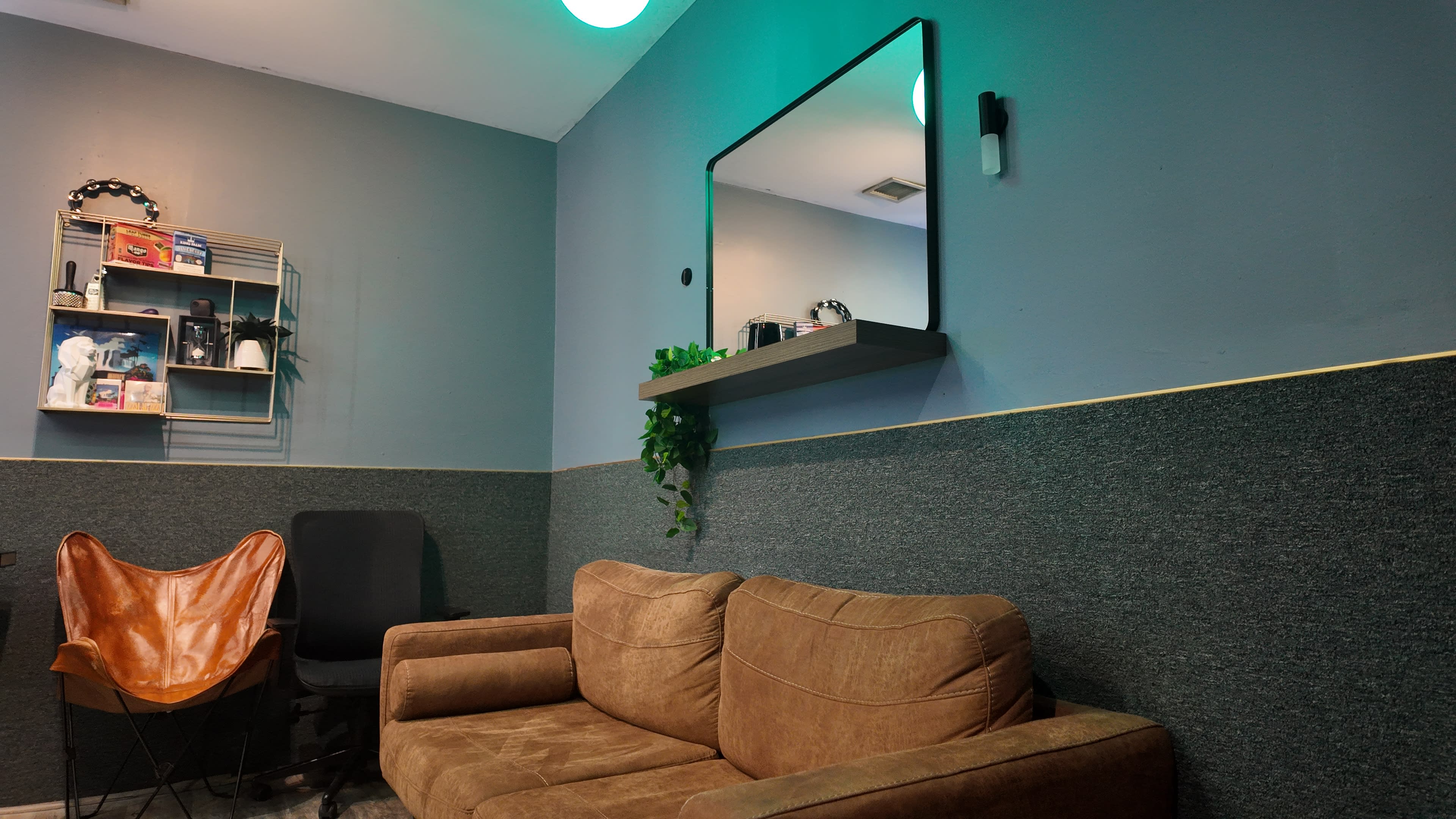 The image shows a cozy room with a brown couch, a black chair, a wall-mounted mirror, and a shelf with decorative items.