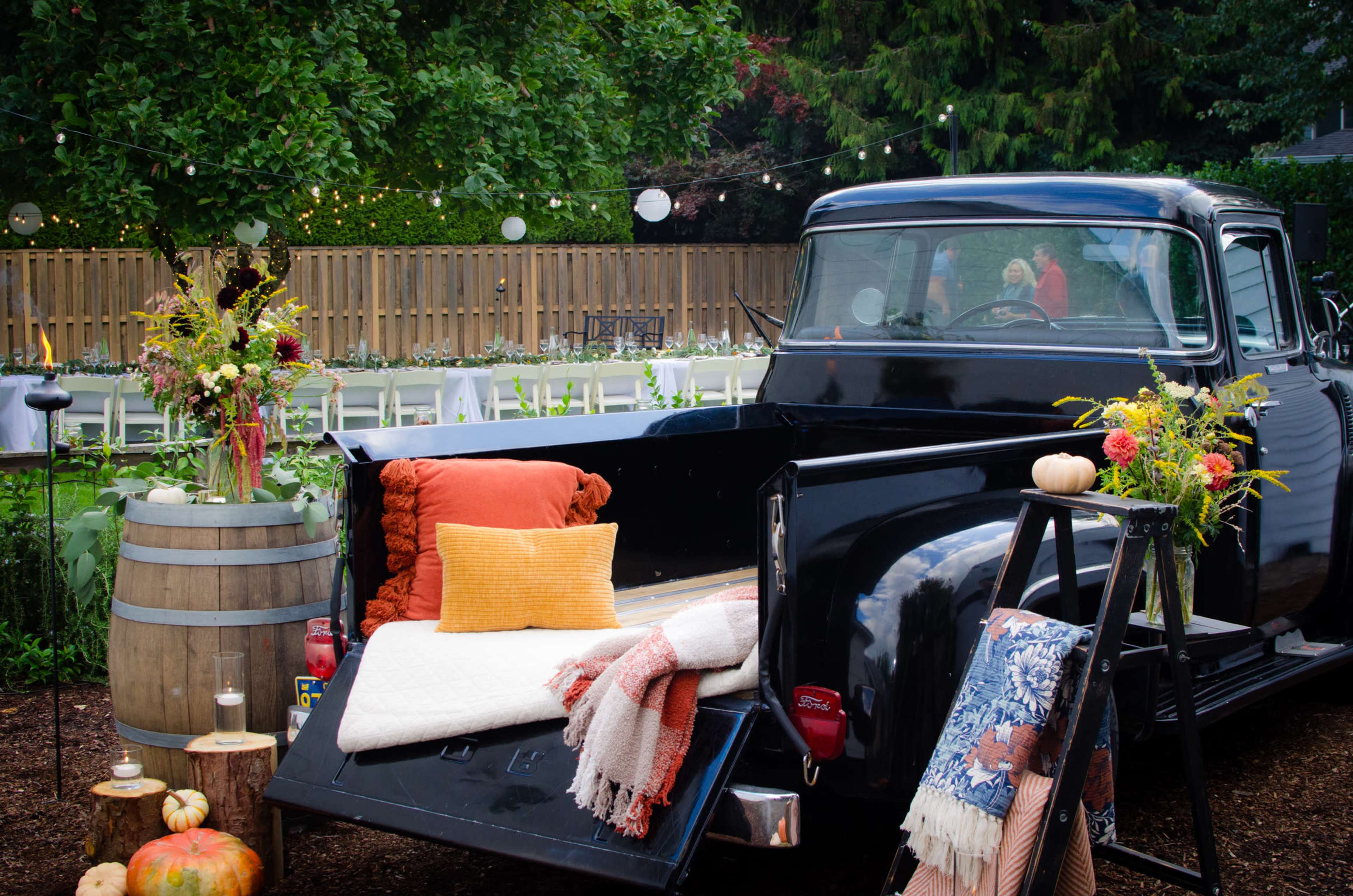 A vintage black pickup truck is parked in a garden setting, featuring colorful cushions and blankets in the bed, with decorative flowers and a wooden barrel nearby.