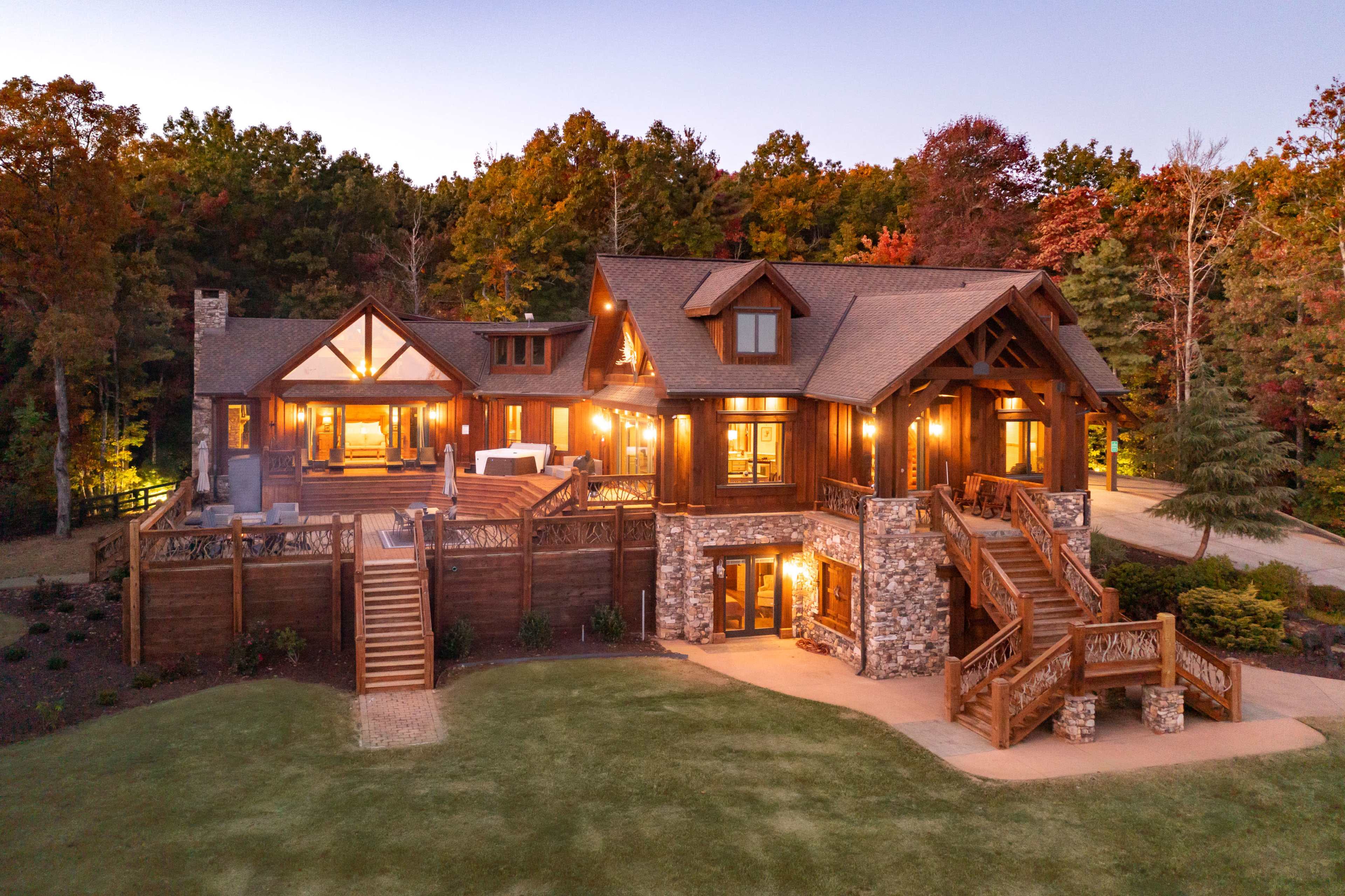 A large, multi-story wooden house with stone accents, surrounded by trees with autumn foliage and lit warmly in the evening.